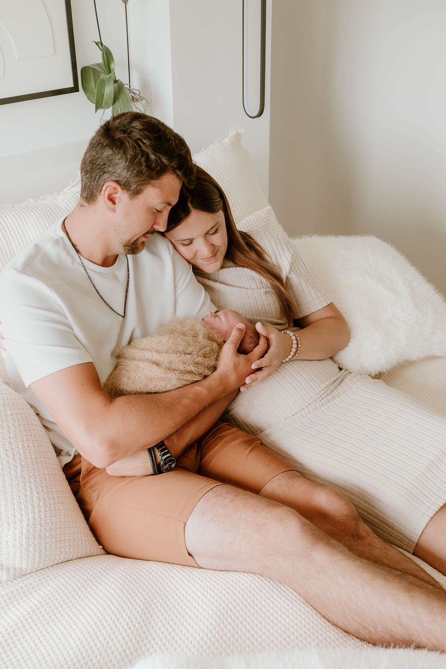A couple sits on a white bed, tenderly cradling their newborn baby in a cozy, softly lit bedroom.