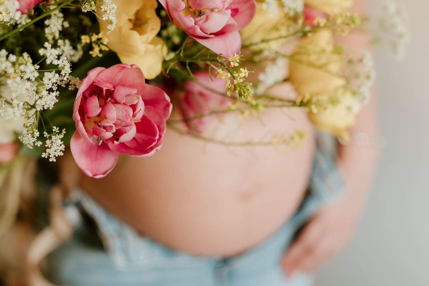 Pregnant woman holding colorful pink and yellow flowers over her bare baby bump in a soft studio maternity photo.