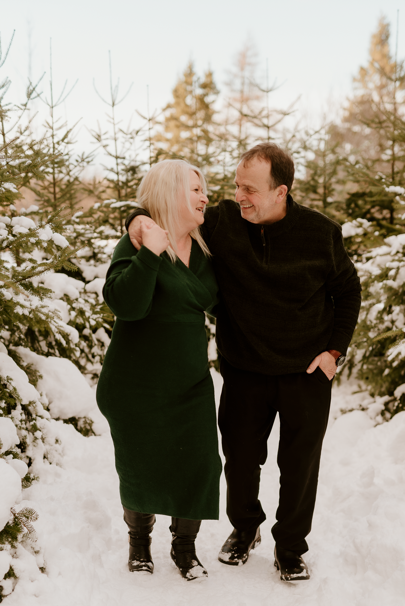 Couple smiling together in snowy Christmas tree farm, woman in green dress and man in black outfit.