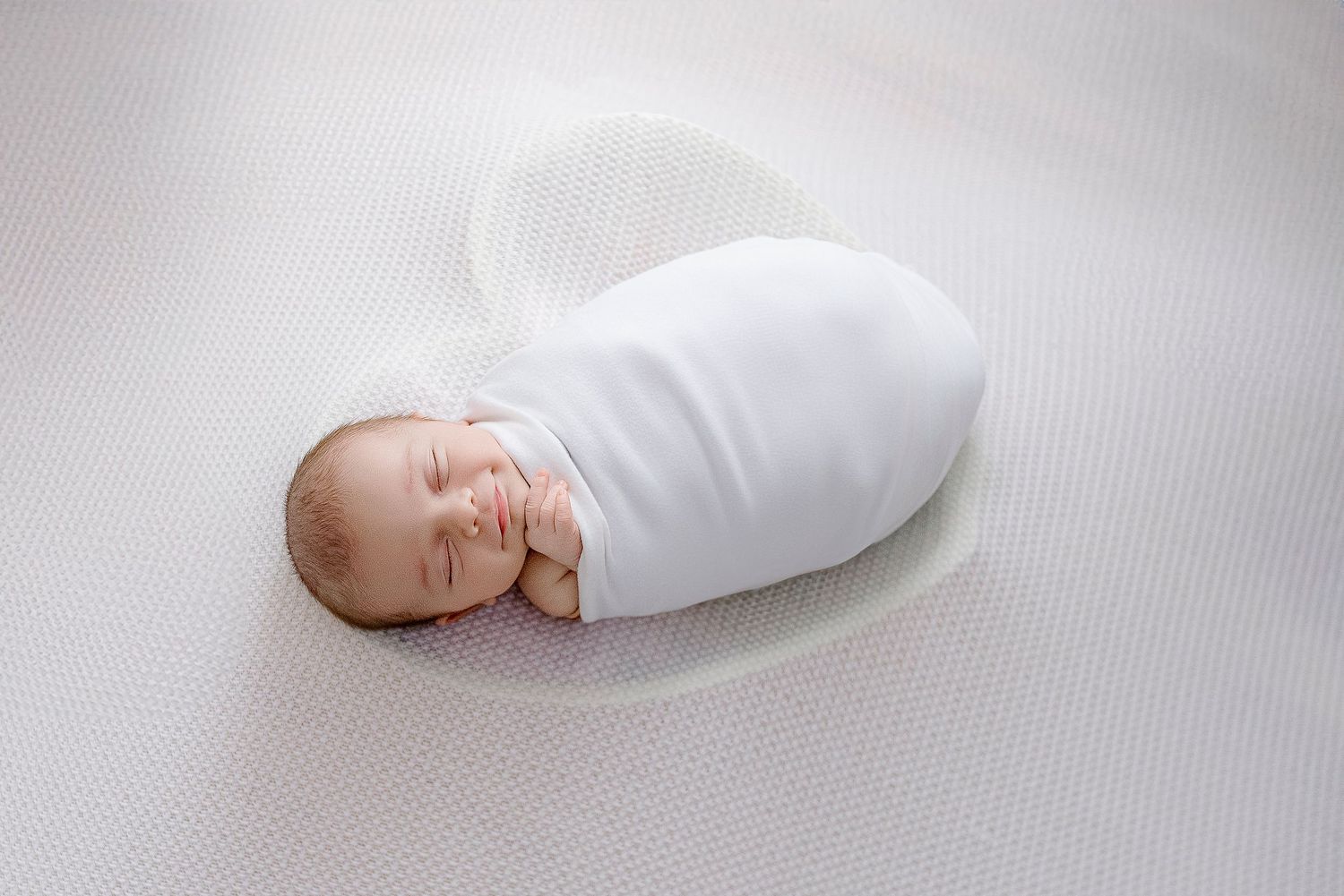 Newborn baby swaddled in white wrap sleeping peacefully on a white textured surface.