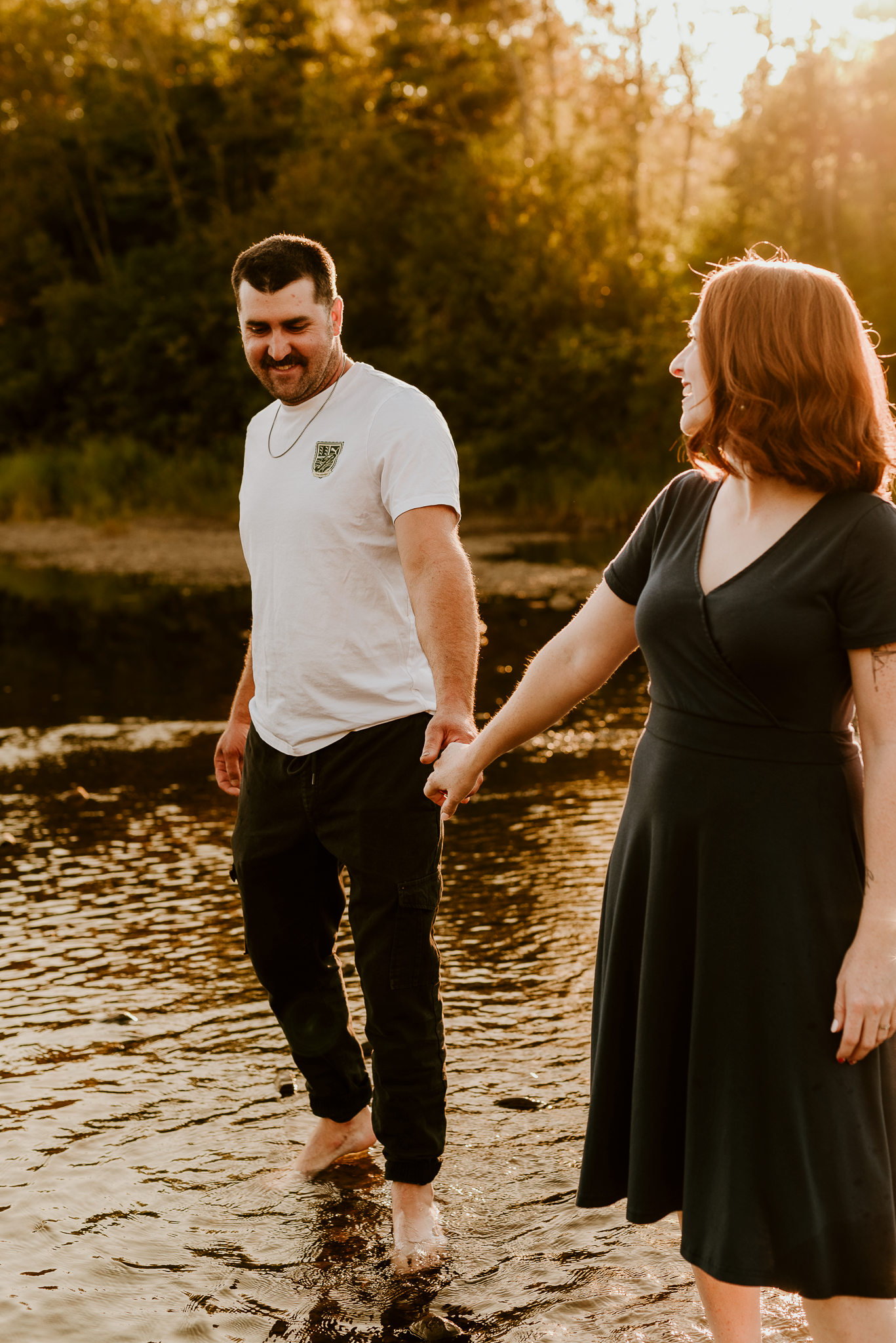 Couple holding hands and laughing while wading in a shallow river at golden hour sunset.