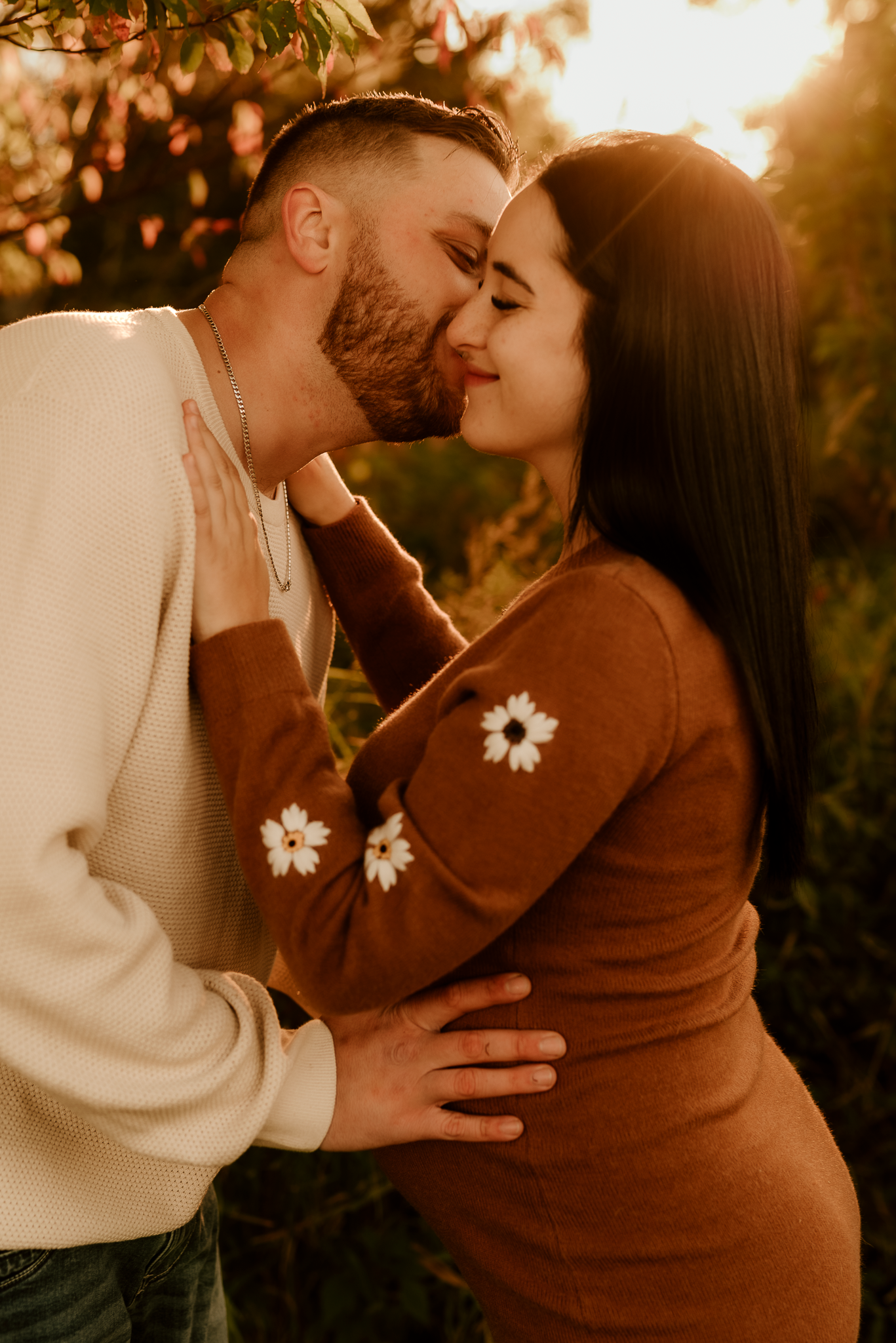 A couple shares a tender kiss outdoors at golden hour, woman in brown floral dress, man in cream sweater.