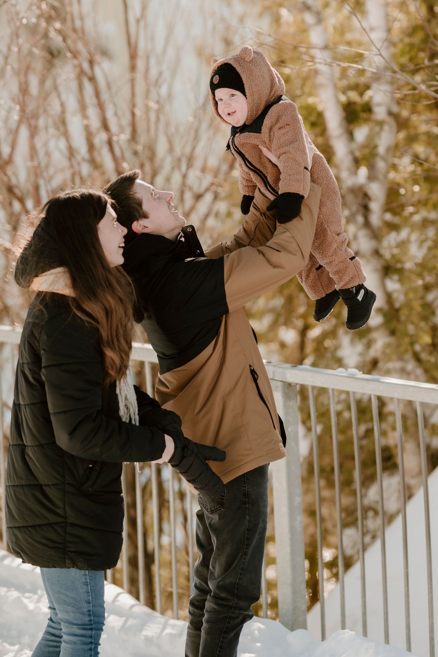 Joyful winter family moment with parents lifting child in matching coats against snowy background.