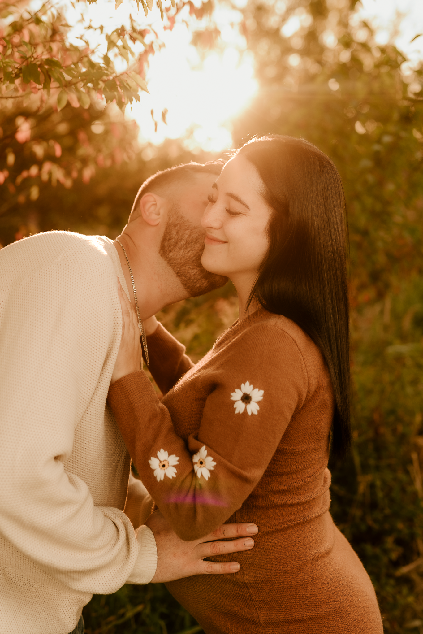 Couple sharing a romantic moment outdoors at golden hour, woman in brown floral sweater, man in white sweatshirt.