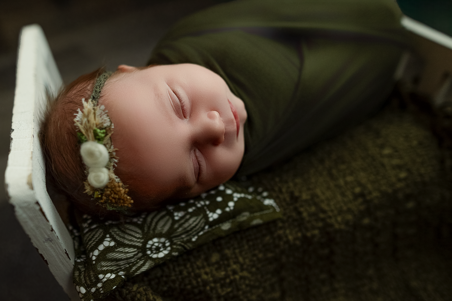Sleeping newborn baby swaddled in green wrap with floral headband resting in a white wooden bed.