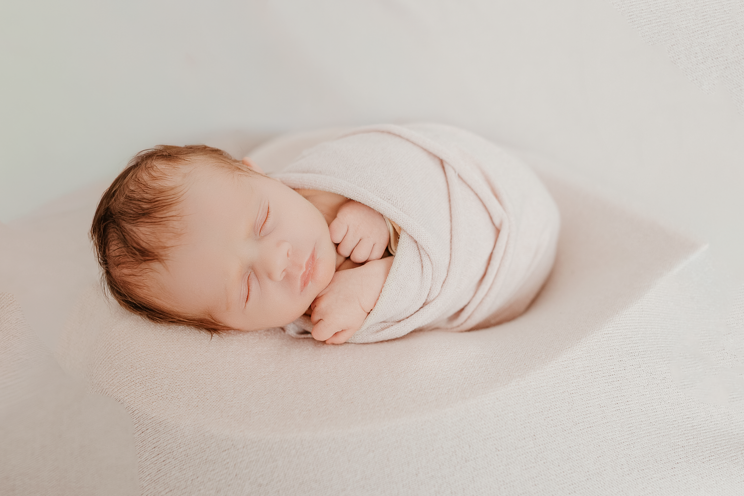 Newborn baby swaddled in white wrap sleeping peacefully on a soft white blanket in a studio setting.