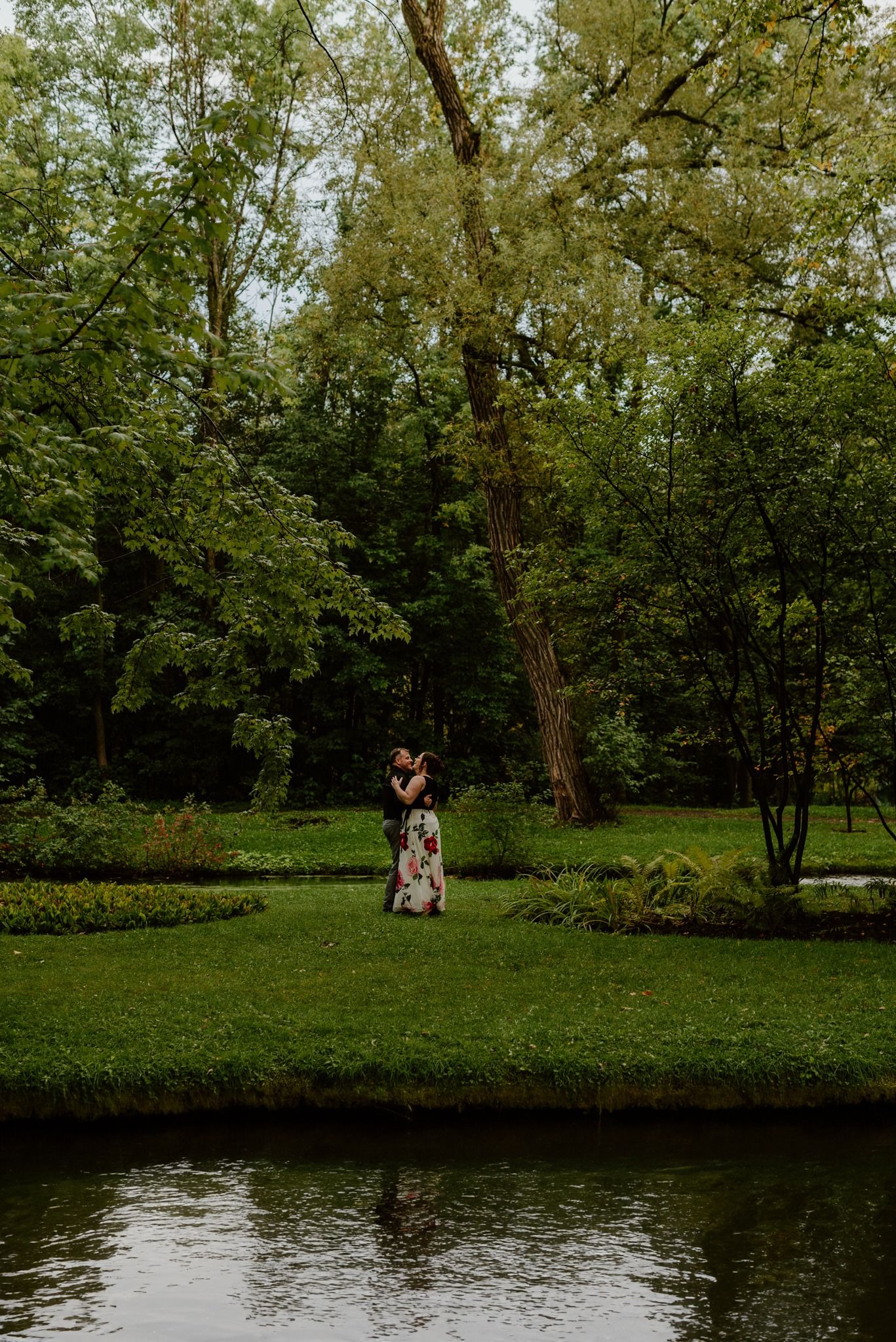 Romantic moment by a pond surrounded by lush green trees and manicured gardens.