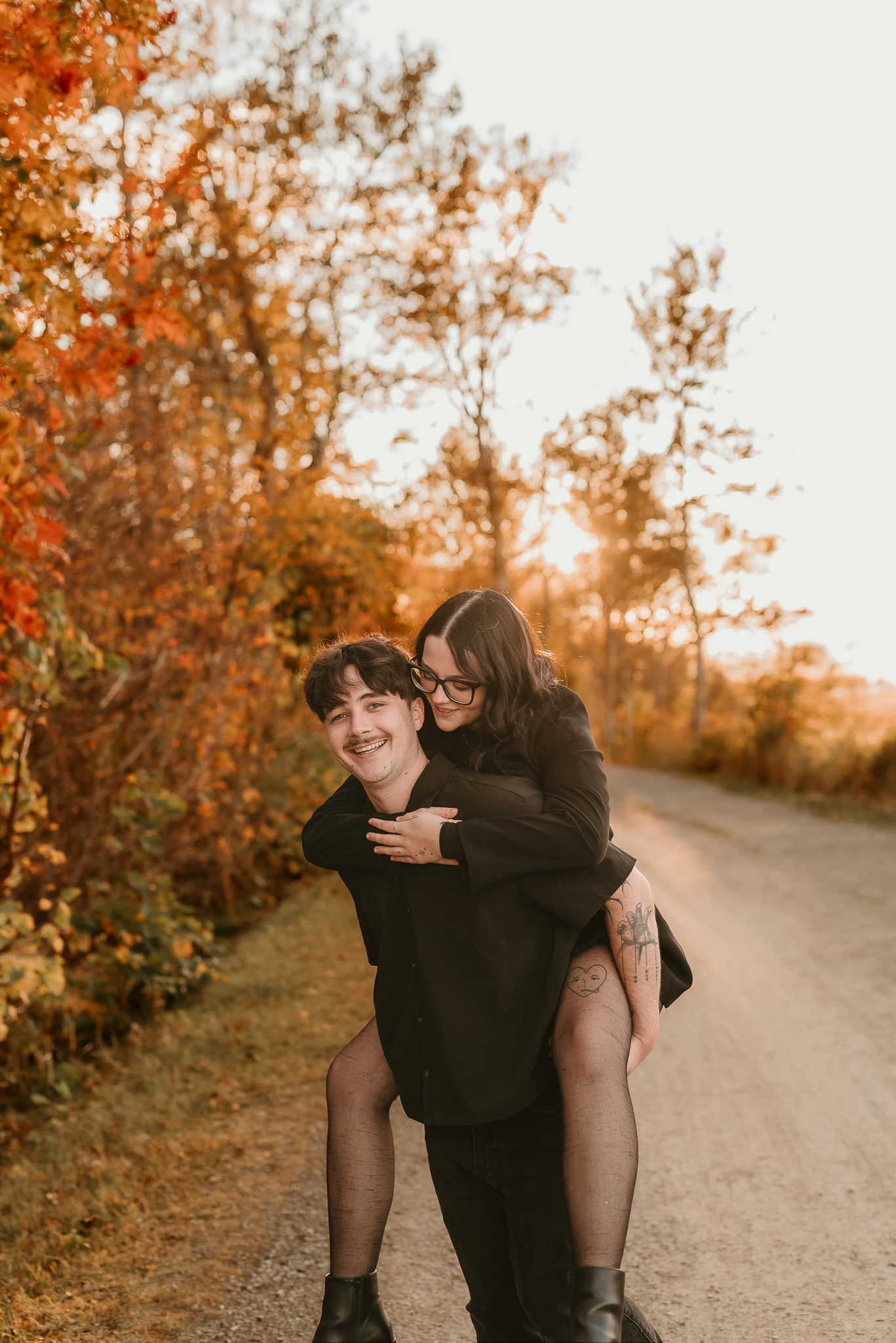 Couple in black outfits sharing a piggyback ride on a dirt path surrounded by vibrant autumn foliage at golden hour.