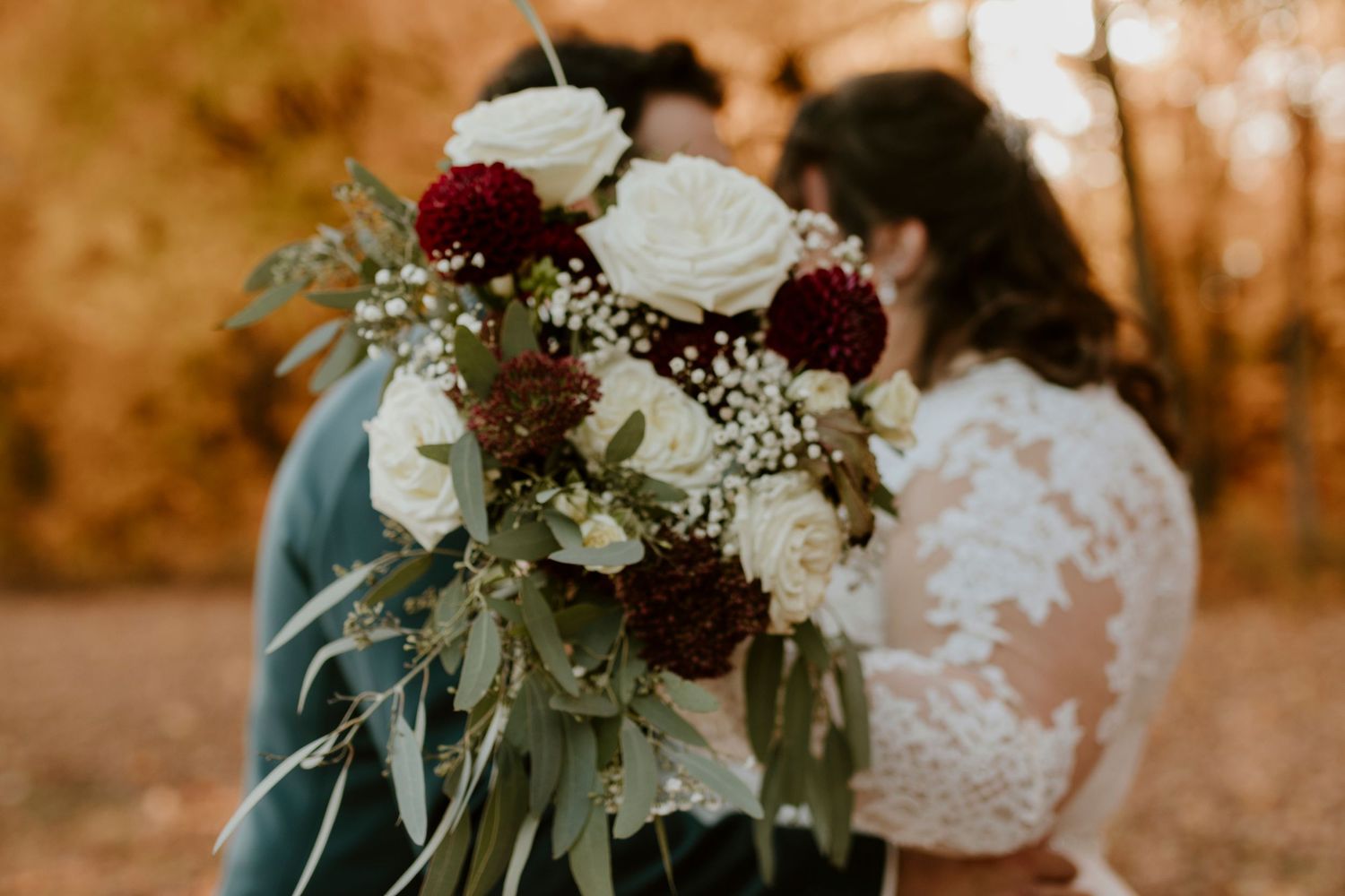 A bride in lace dress holds a bouquet of white roses and dark red dahlias with greenery at an autumn wedding.