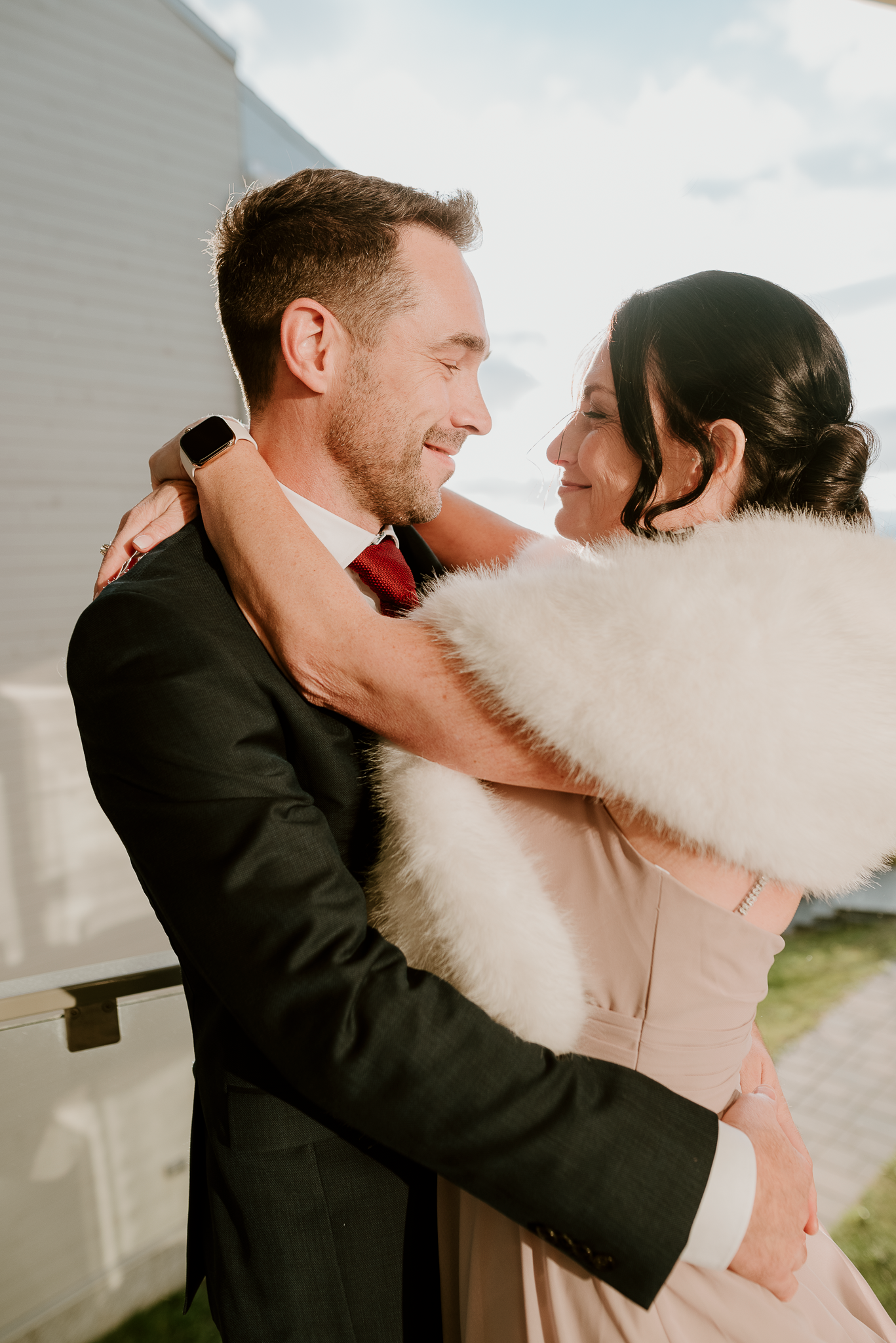 Elegant couple embracing outdoors, bride wearing fur shawl with groom in formal black suit.