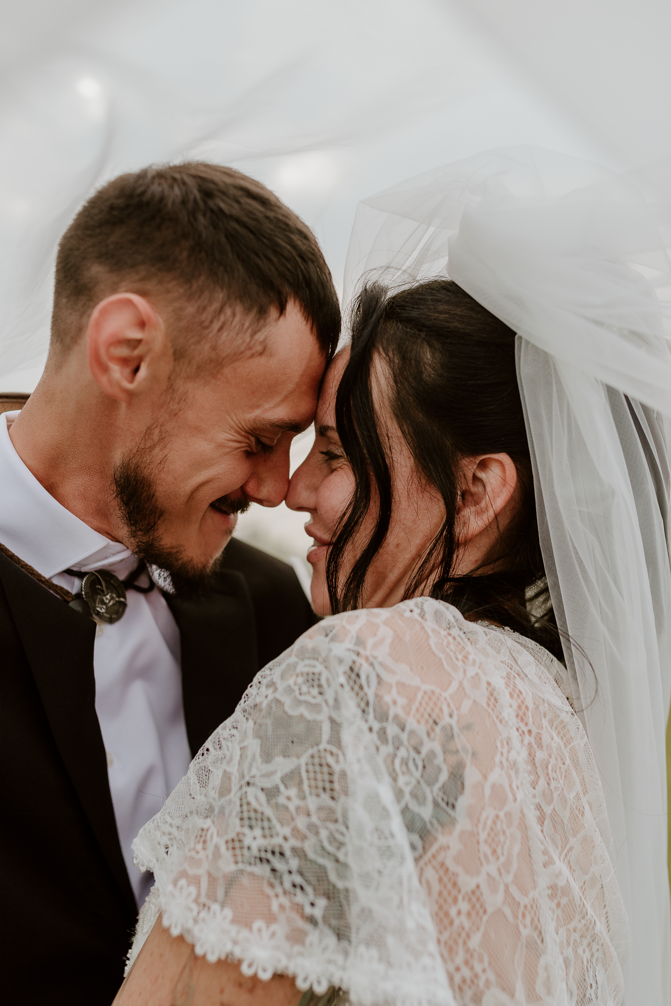 A bride and groom share an intimate forehead-to-forehead moment under a flowing white veil on their wedding day.