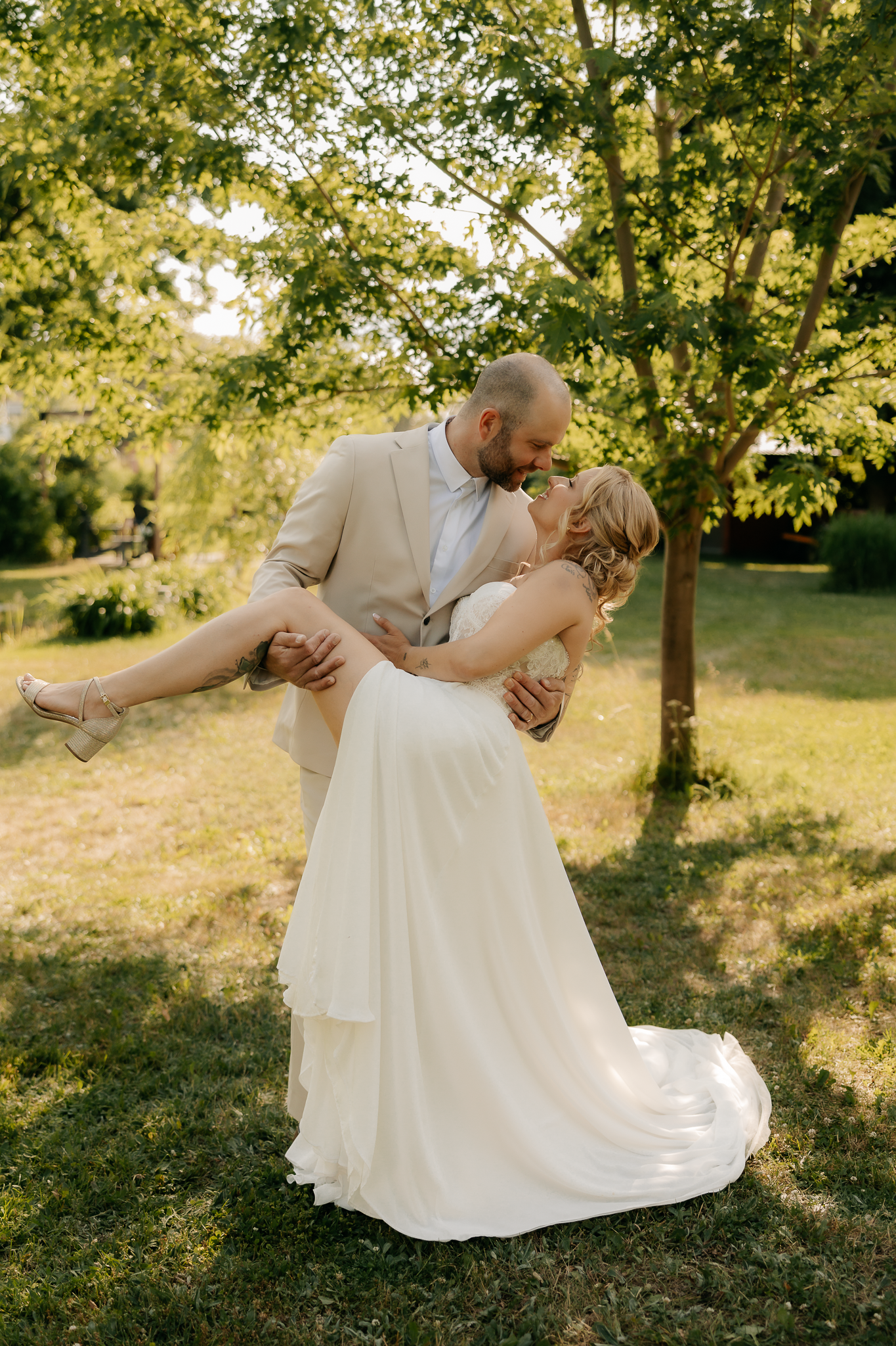 Groom in tan suit lifts bride in white gown for a romantic kiss outdoors in a sunlit garden.