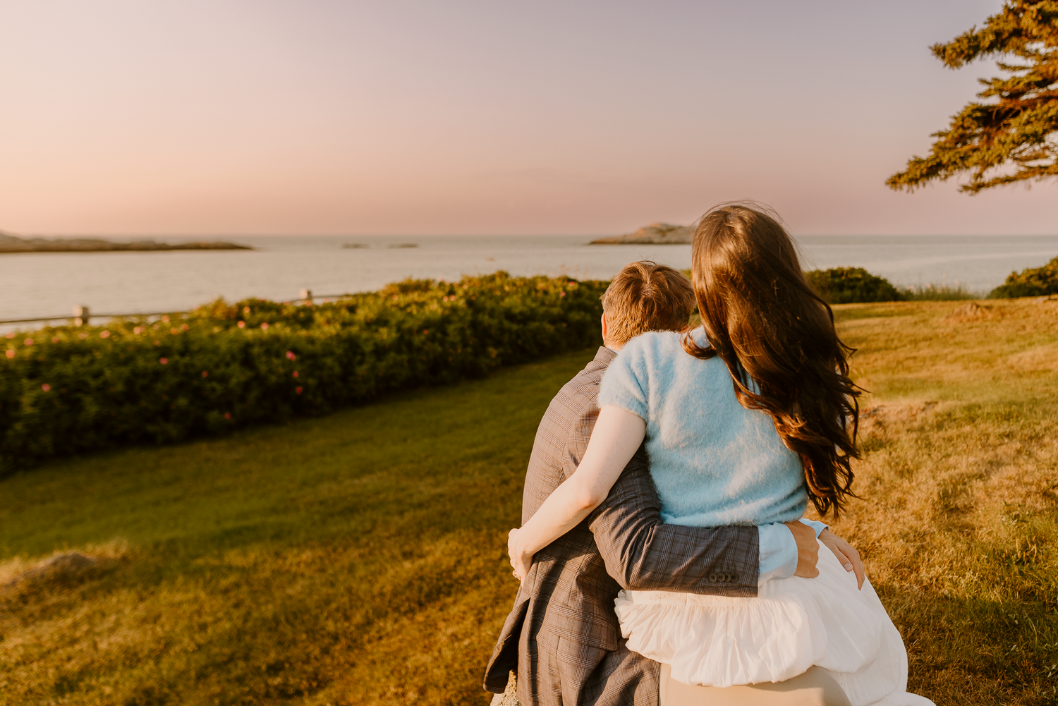 A mother and child embrace while gazing at a serene coastal sunset over lush green hills.