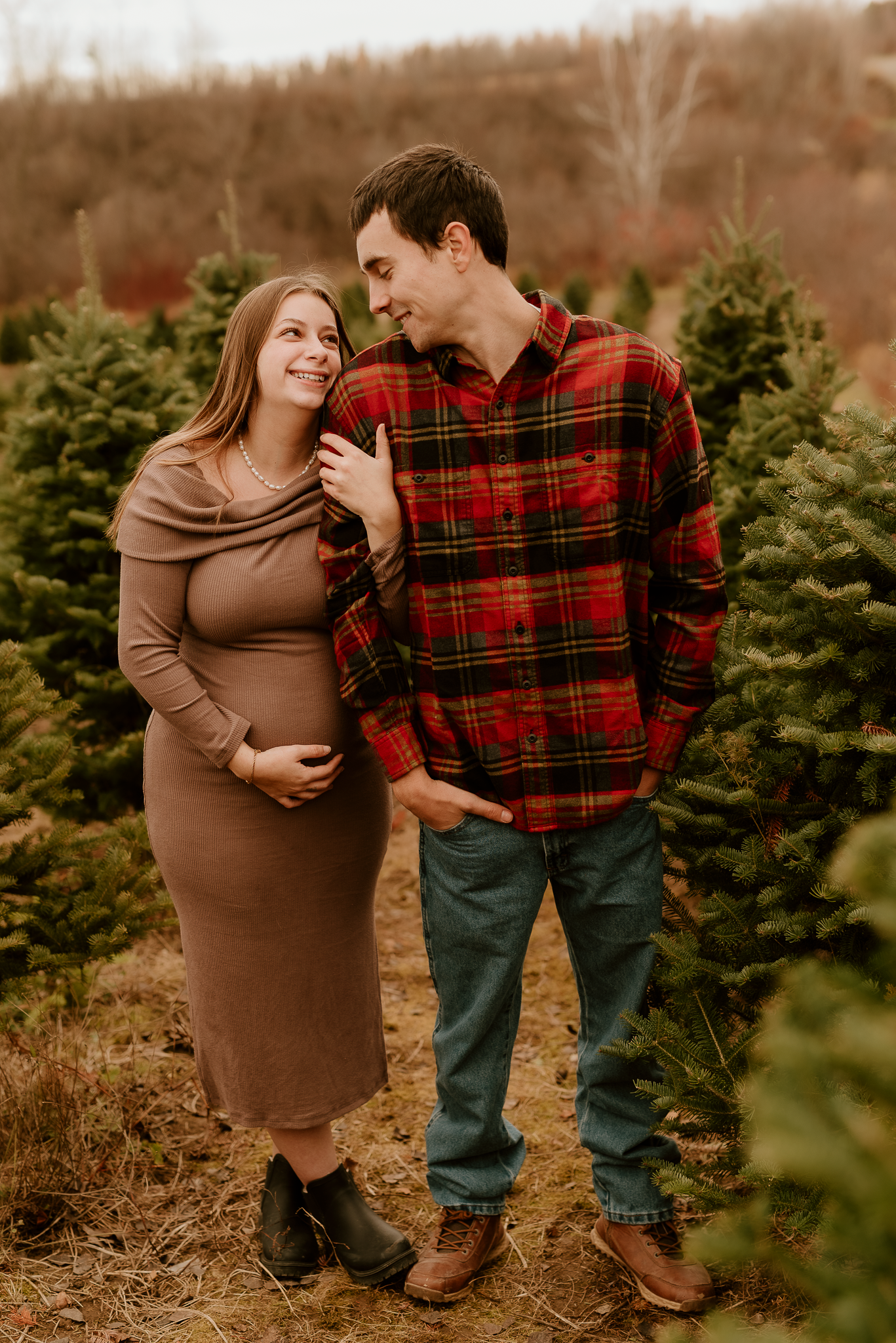 Pregnant couple smiling at each other at a Christmas tree farm, woman in brown dress, man in red plaid flannel shirt.