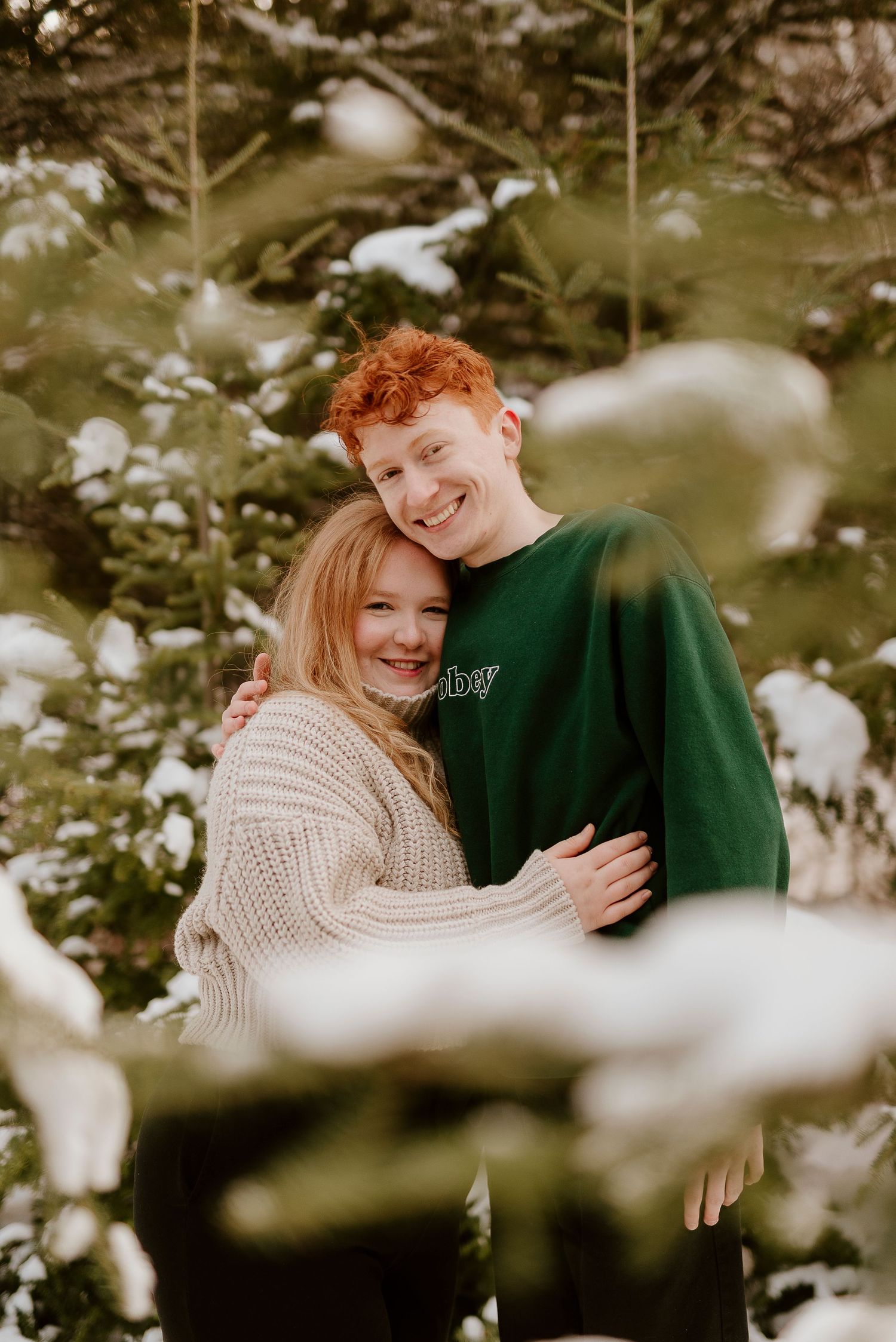A smiling couple embraces outdoors among snow-dusted evergreen trees, the man in a green sweatshirt hugging a woman in a cream sweater.