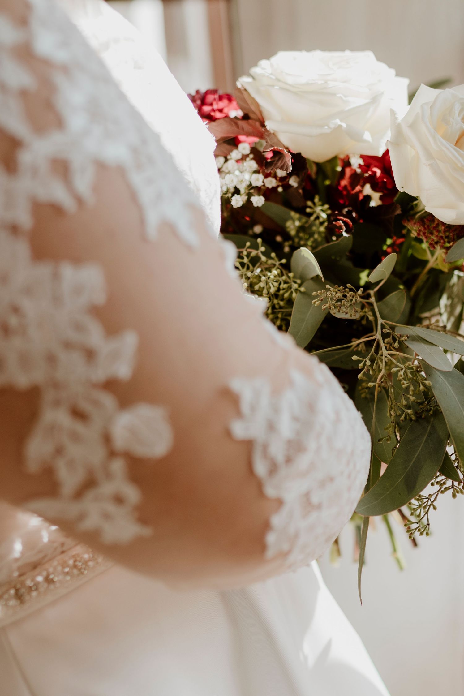 Bride in lace wedding dress holding a bouquet of white roses and red flowers with greenery.
