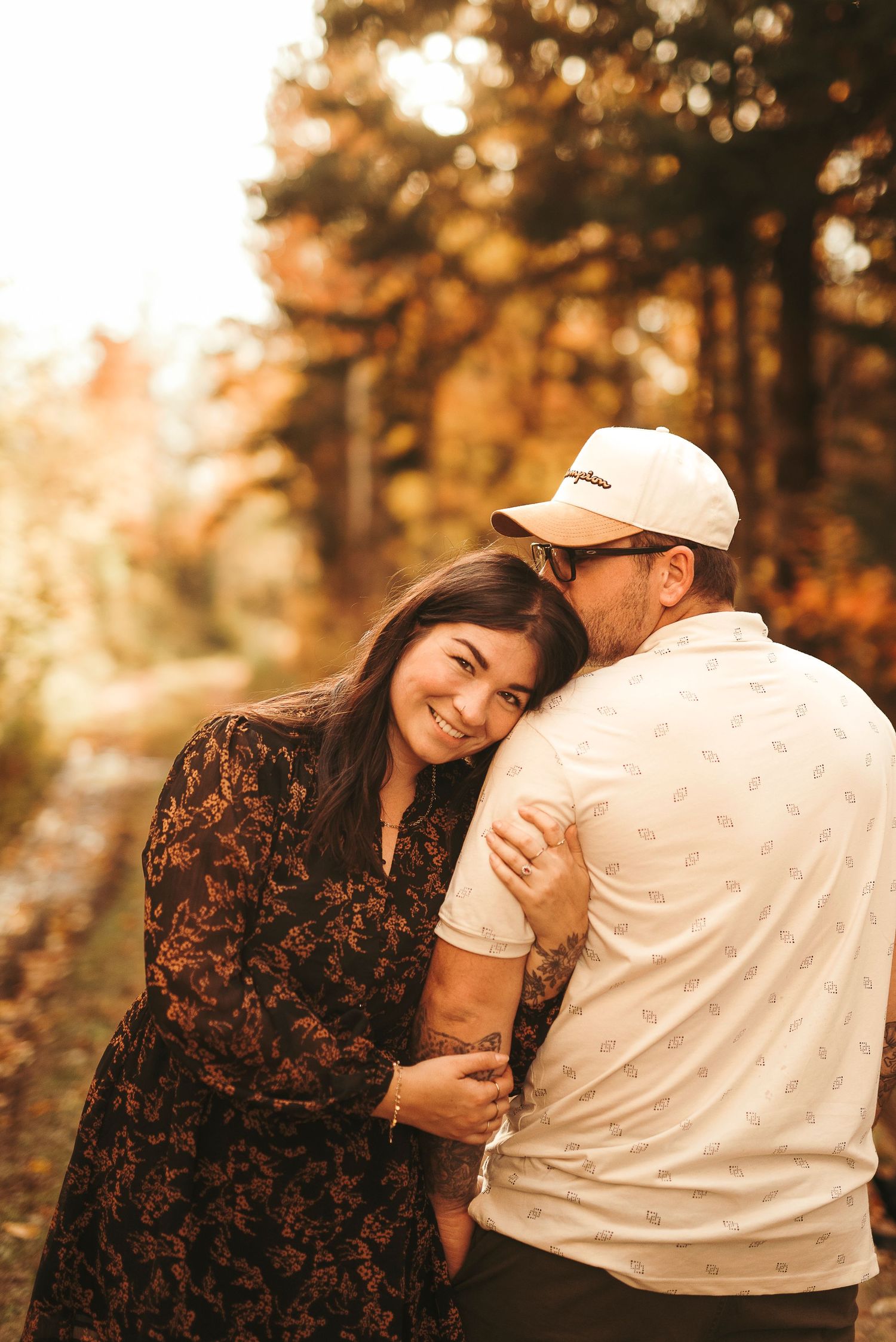 Couple embracing outdoors in autumn forest, woman smiling in floral dress, man in white cap and shirt, warm golden fall foliage.