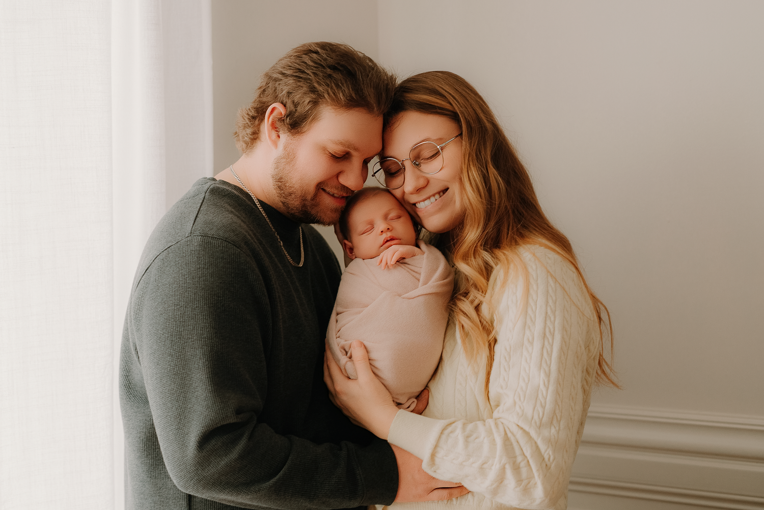 Happy parents holding their swaddled newborn baby, sharing a tender family moment in a bright, softly lit room.