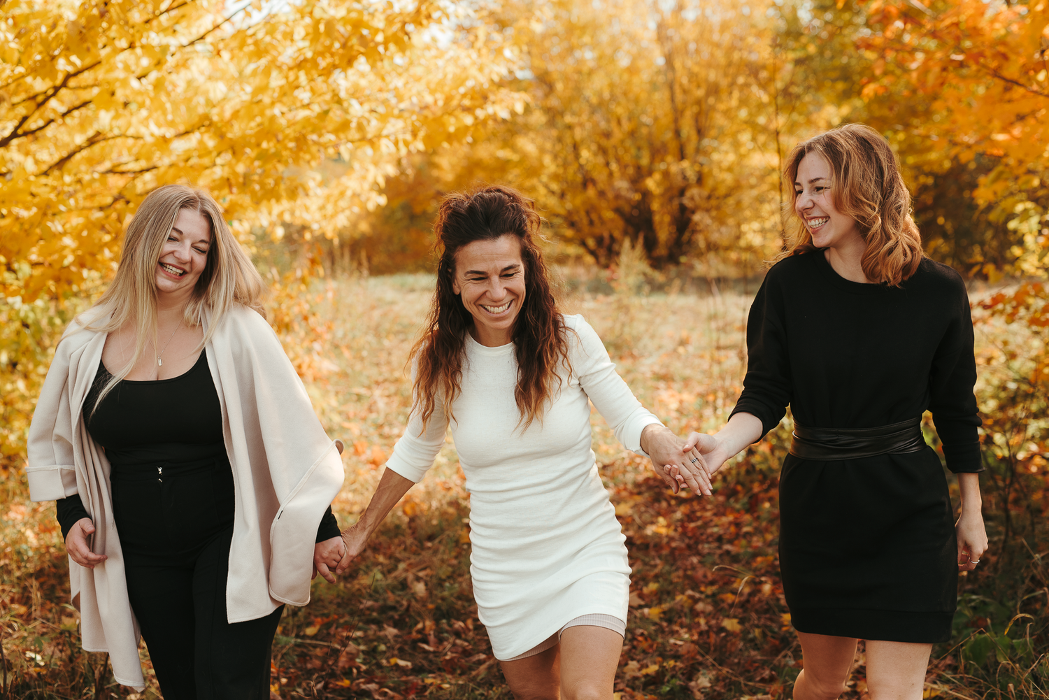 Three women holding hands and laughing while walking through an autumn forest with golden yellow trees.