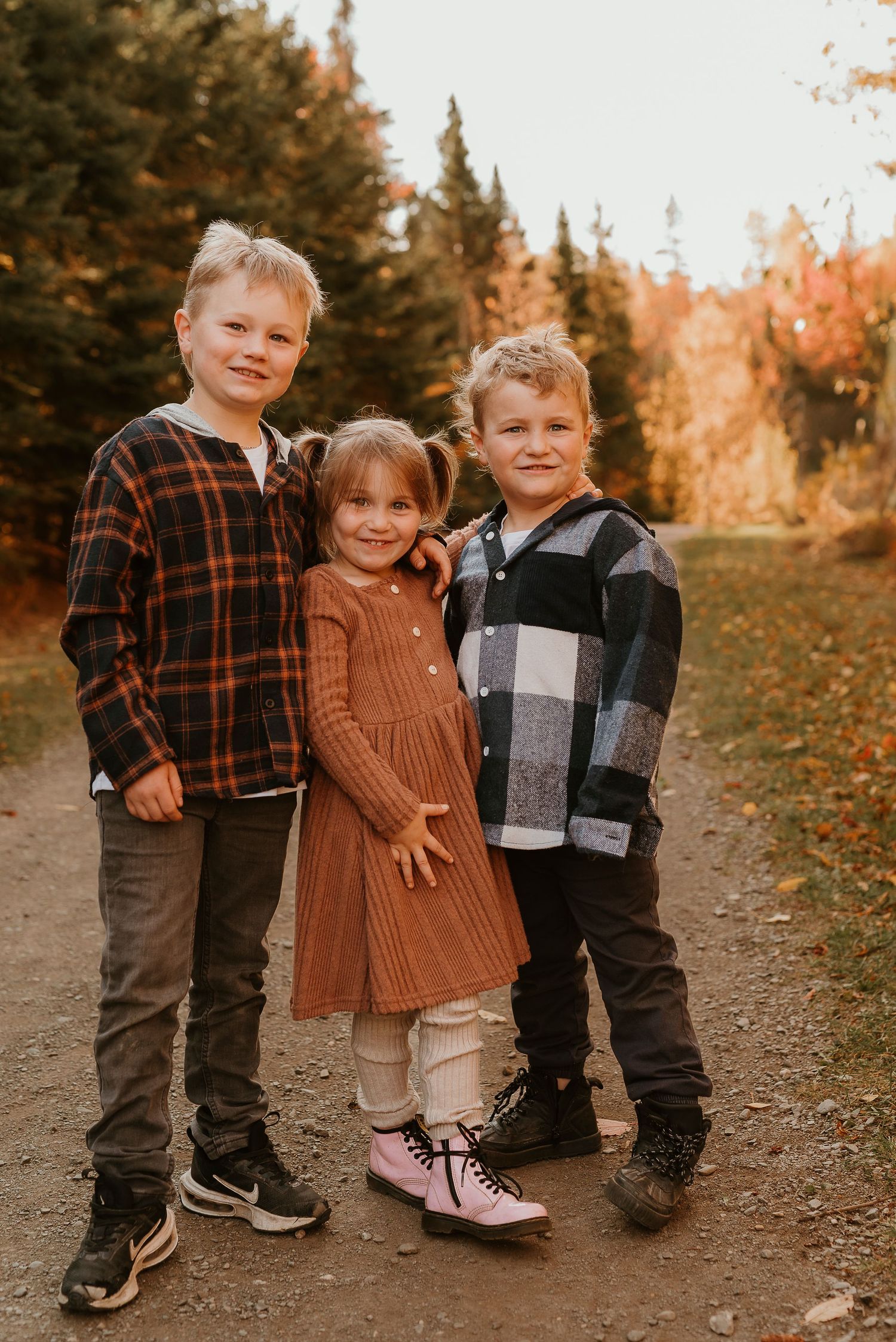 Three children pose on a fall forest path, wearing plaid shirts and a rust dress, surrounded by colorful autumn foliage.
