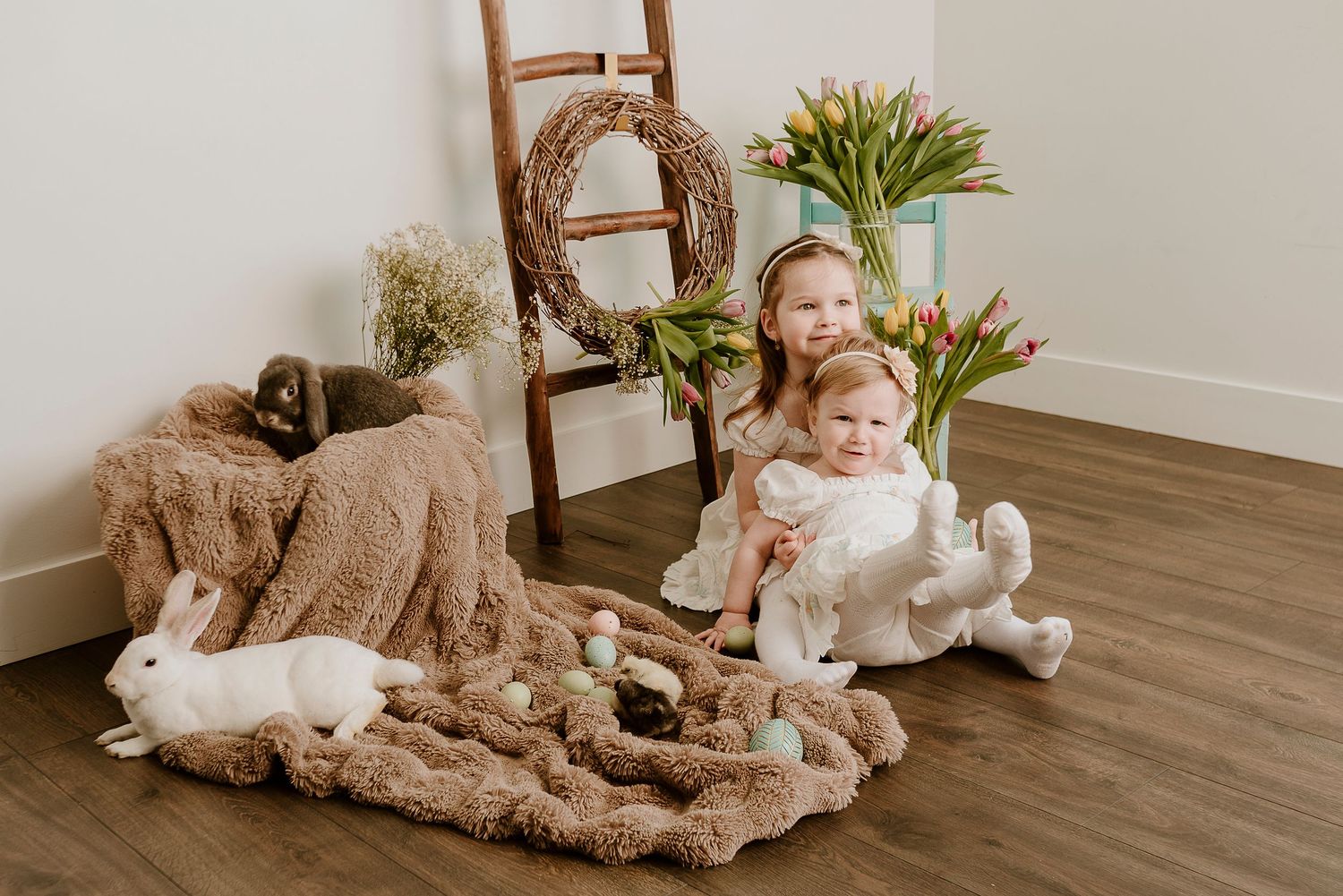Two young girls in white dresses sit on a fluffy blanket with Easter eggs and white bunnies near tulips and a wreath.