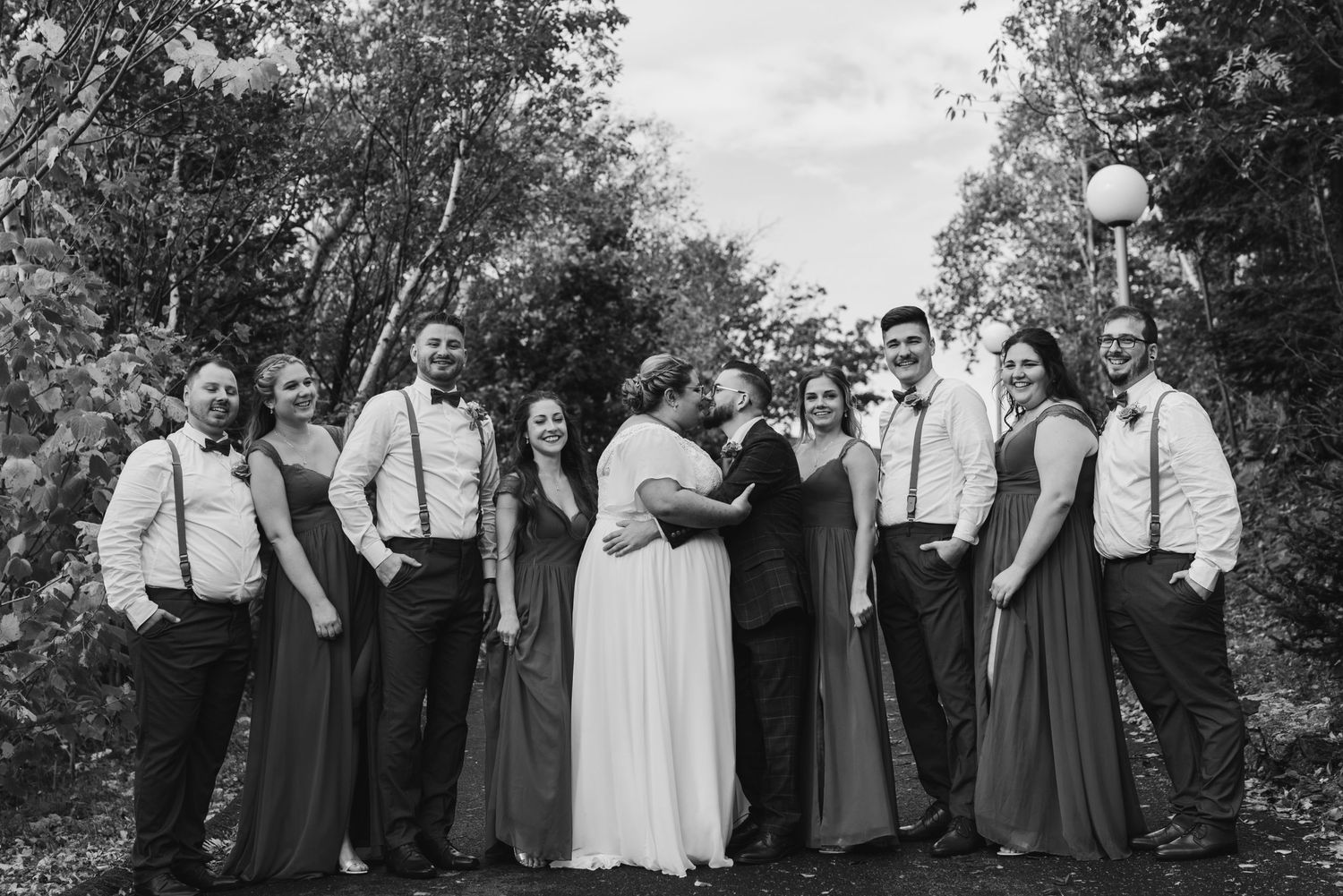 Wedding party in formal attire poses together outdoors under trees.