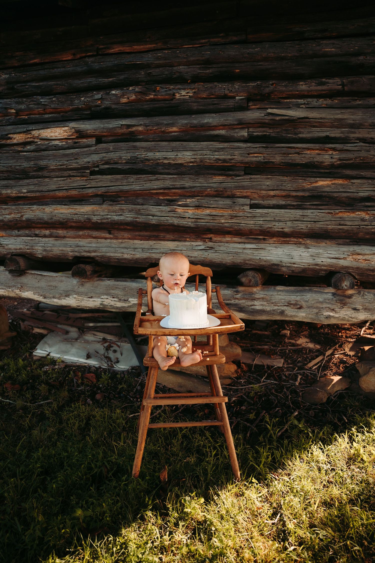 Baby sitting in wooden chair against weathered log cabin wall with sunlit grass.