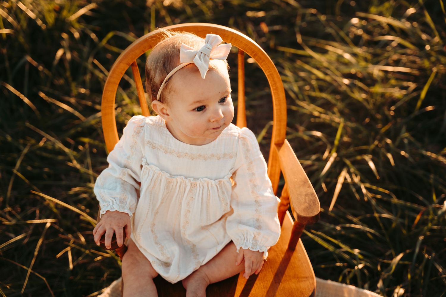 Baby in white dress sitting in wooden chair outdoors with natural lighting.