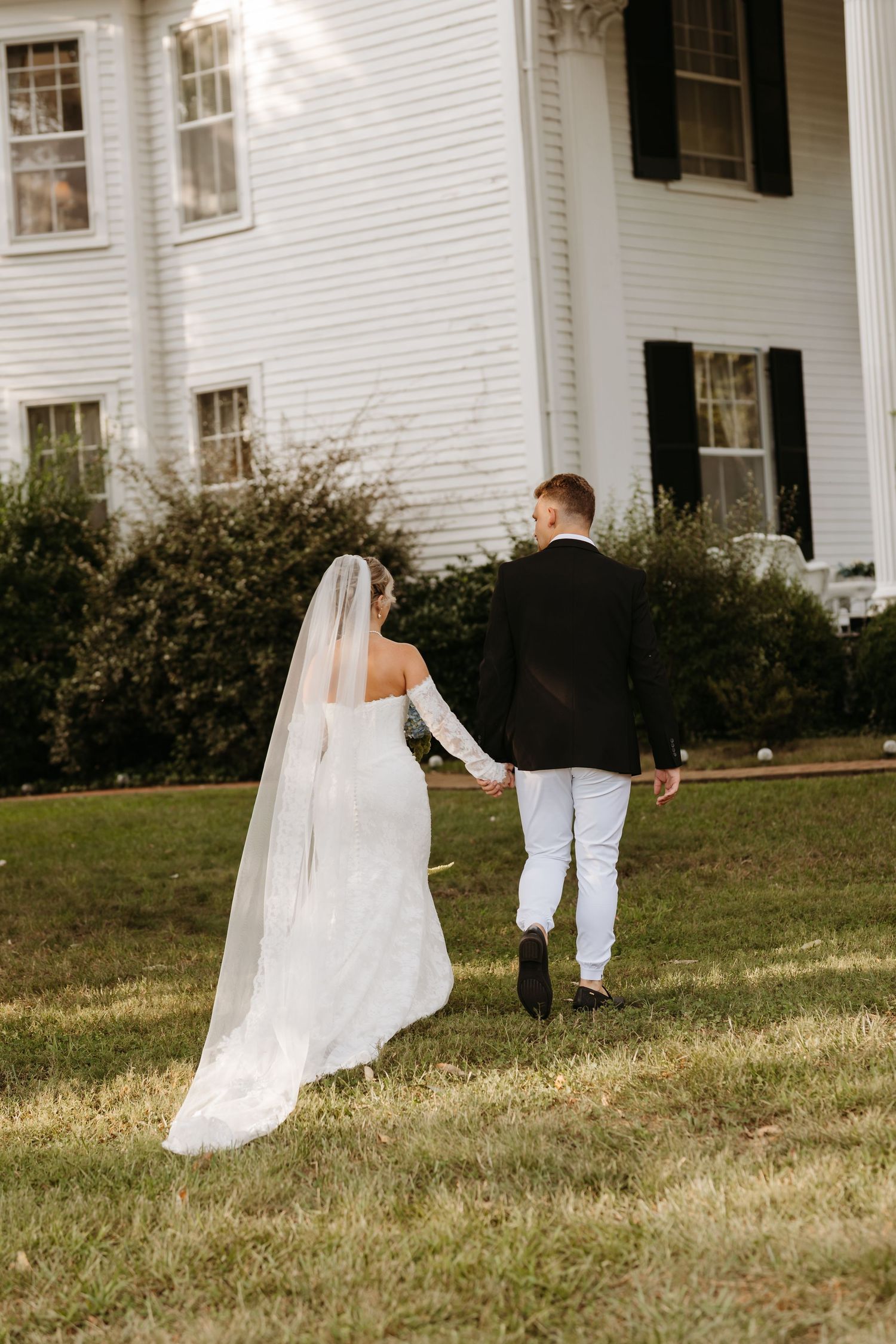A bride in a white gown and veil holds hands with a groom in a black suit walking toward a white colonial home.