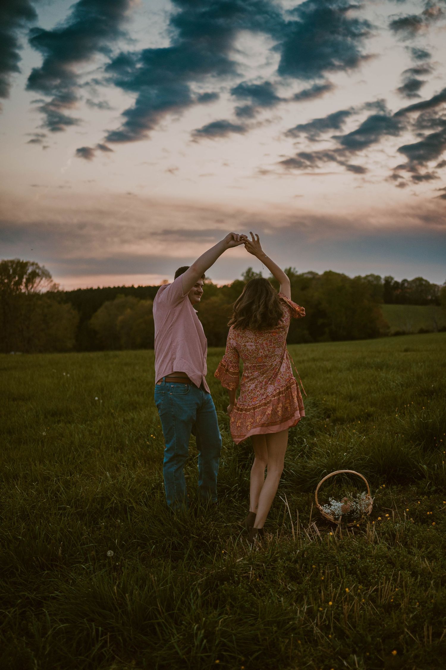 A couple dances in a meadow during golden hour with dramatic clouds in the background.