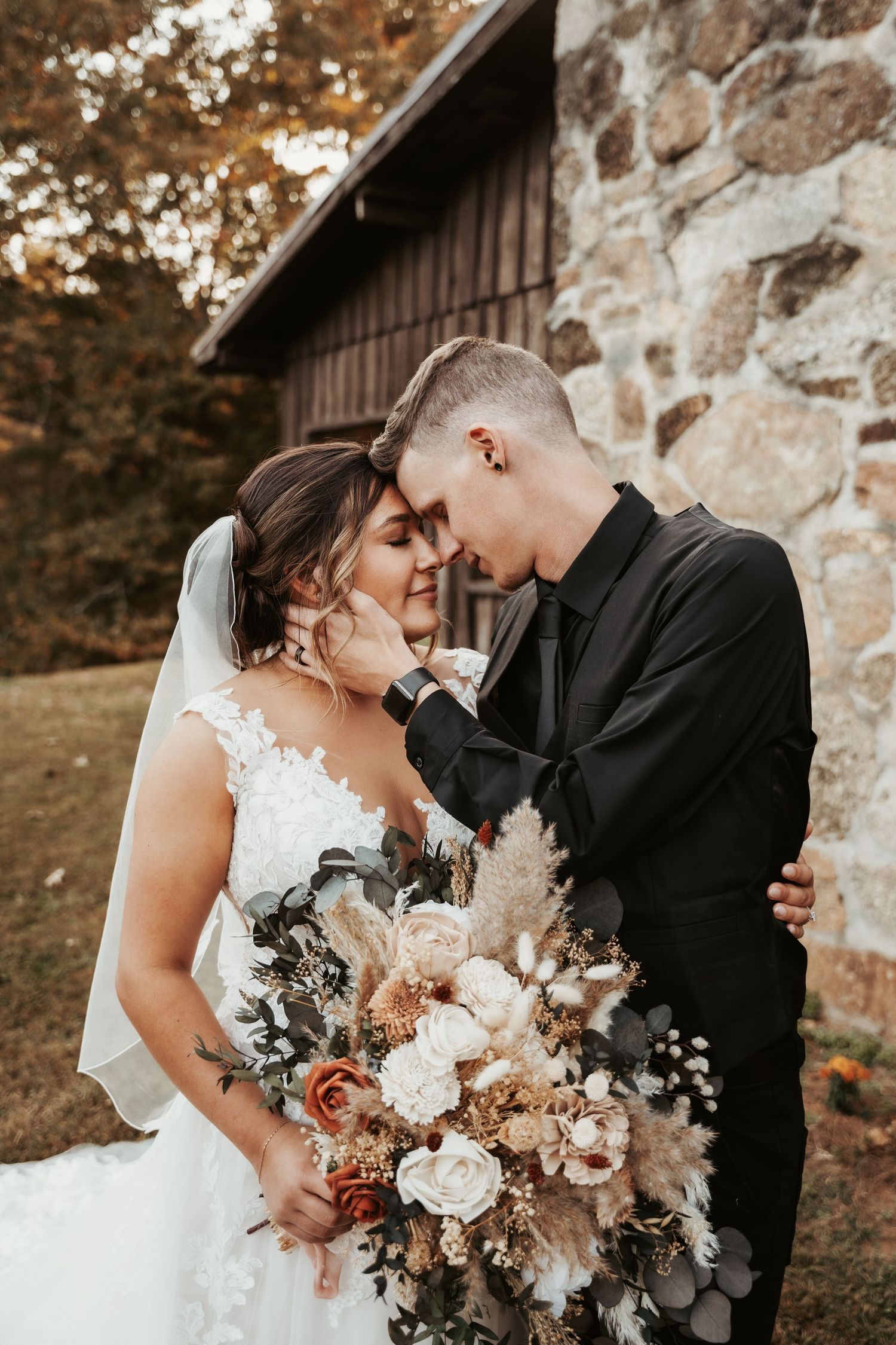 Intimate wedding portrait with couple sharing tender moment beside rustic barn with beautiful bouquet.
