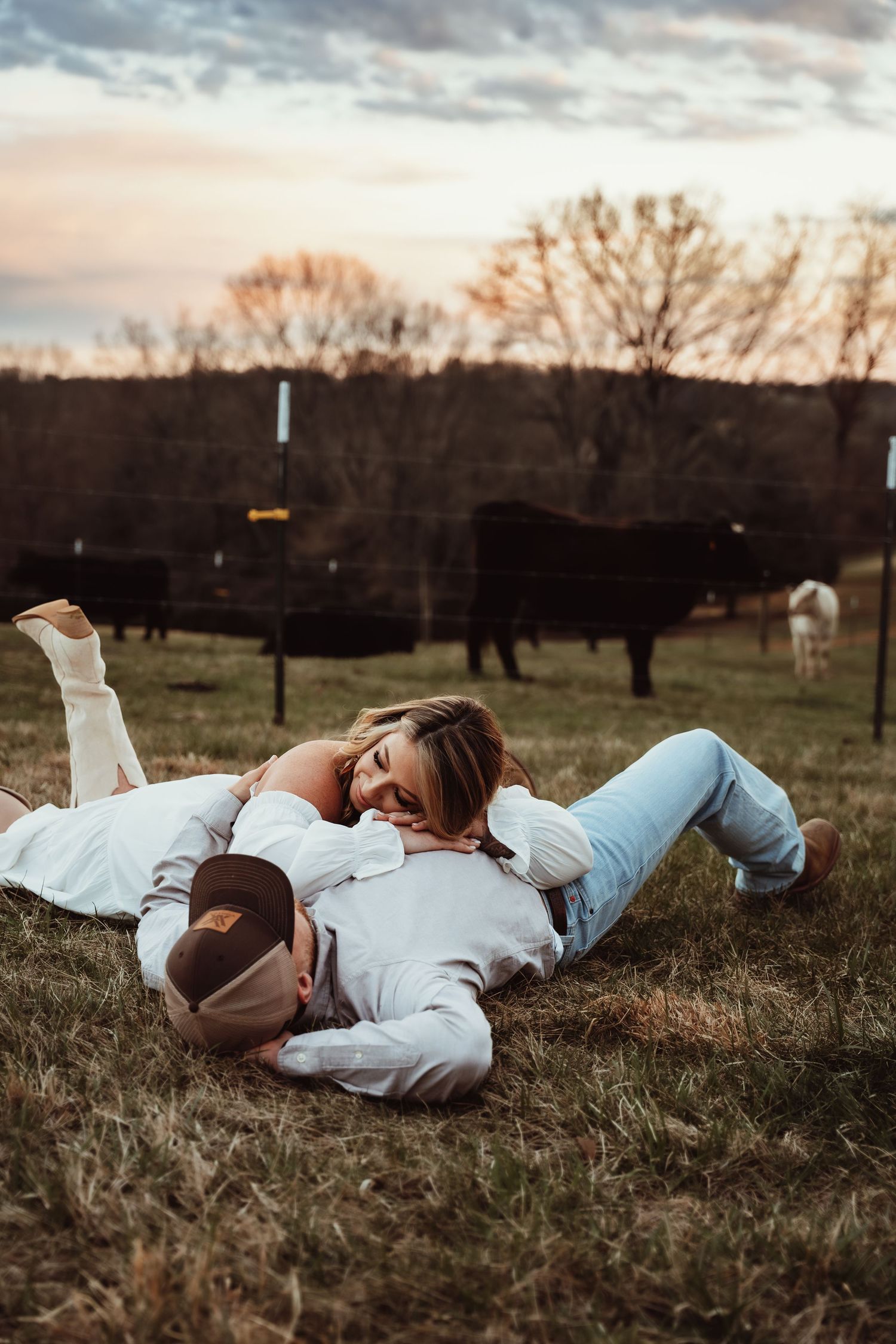 A couple relaxes in casual attire on grass with cattle visible in the distance.
