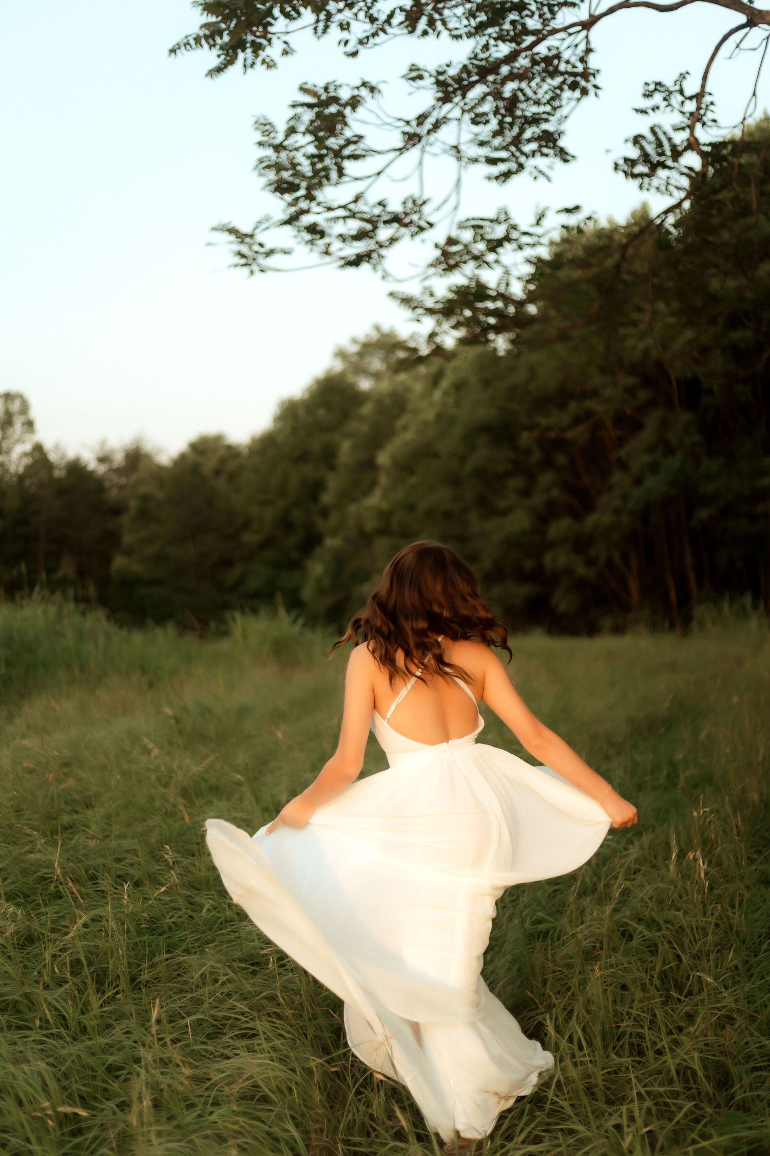Woman in flowing white backless dress running through green meadow with trees at golden hour.