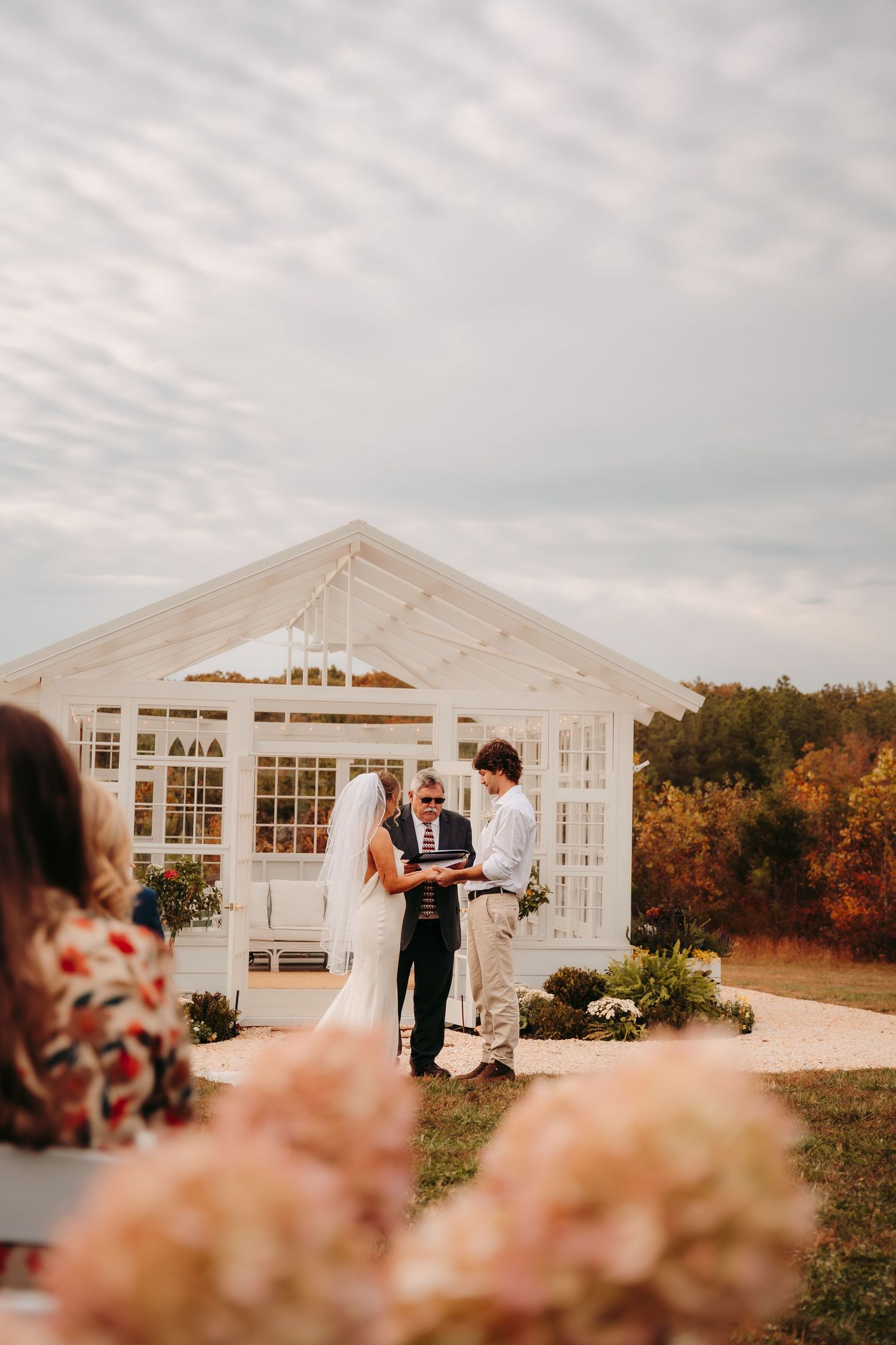 Wedding ceremony taking place in front of a white greenhouse structure with autumn flowers in the foreground.