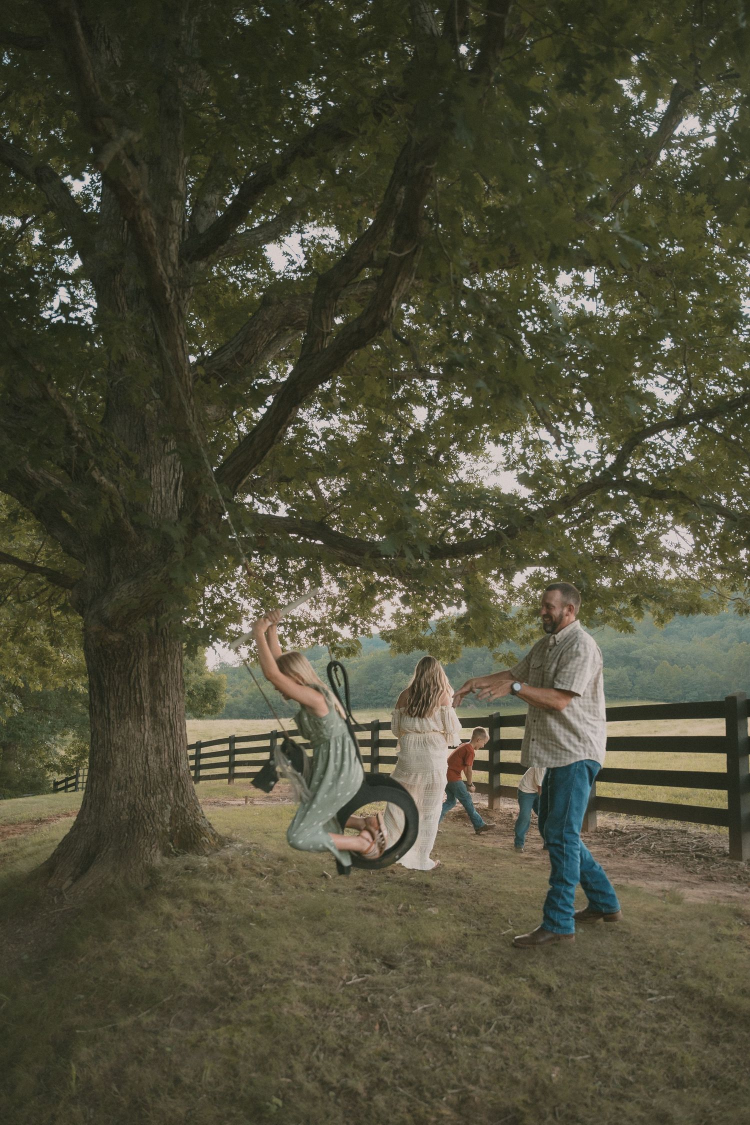 Family plays and runs together near a large tree in a pastoral setting.