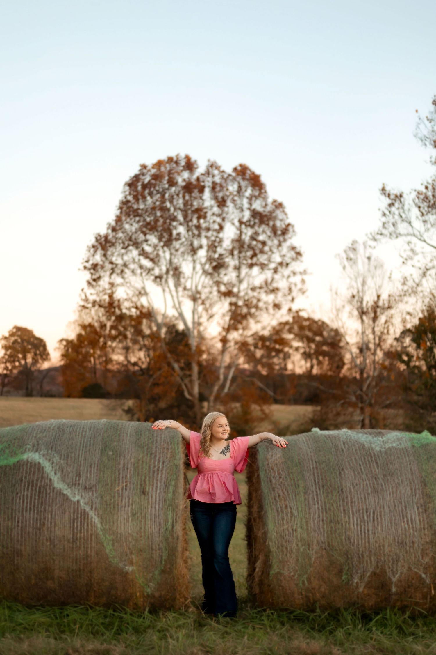 A blonde woman in a pink top and jeans leans against hay bales in a field during golden hour.