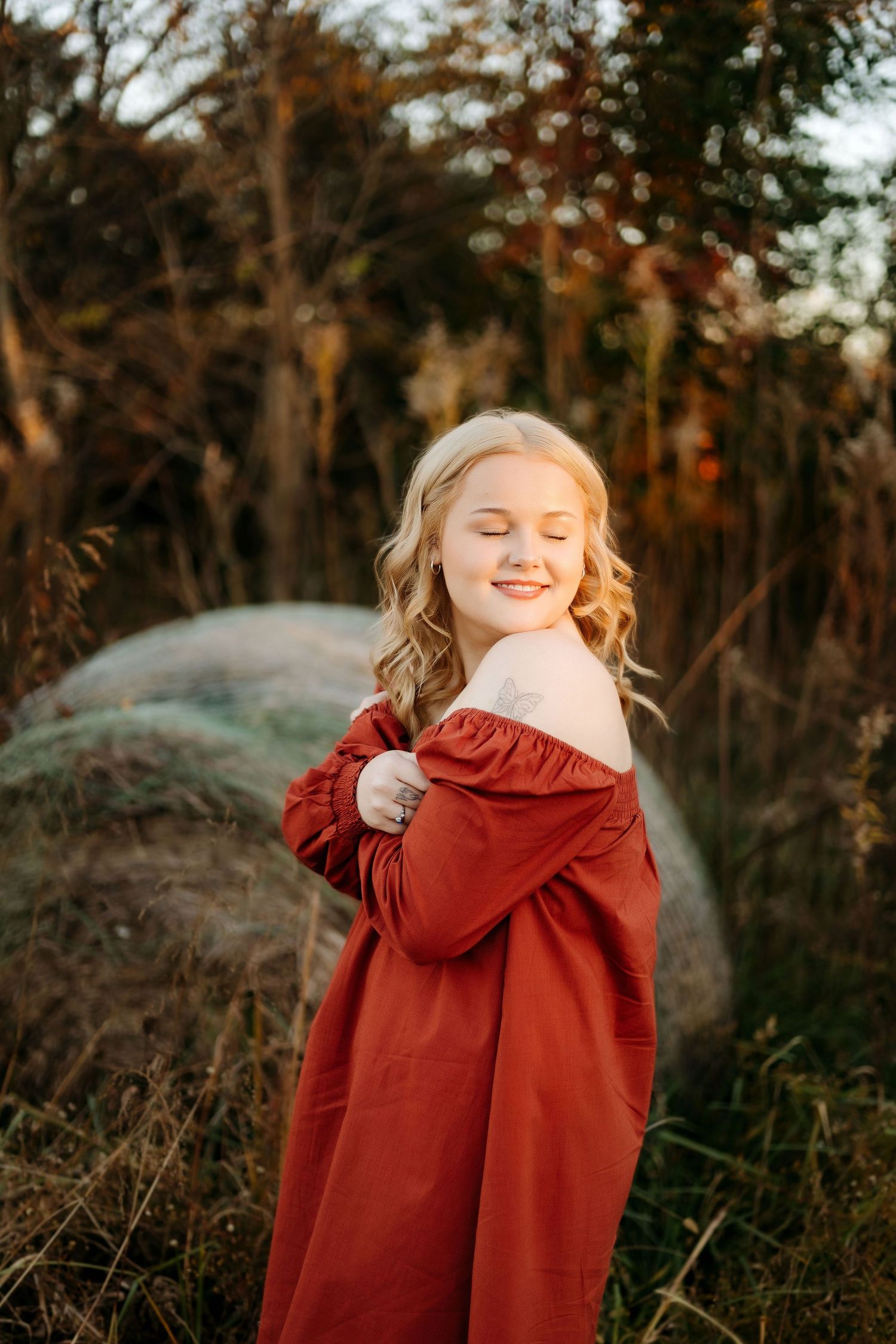 Blonde girl in rust red off-shoulder dress smiling with eyes closed in golden autumn field.