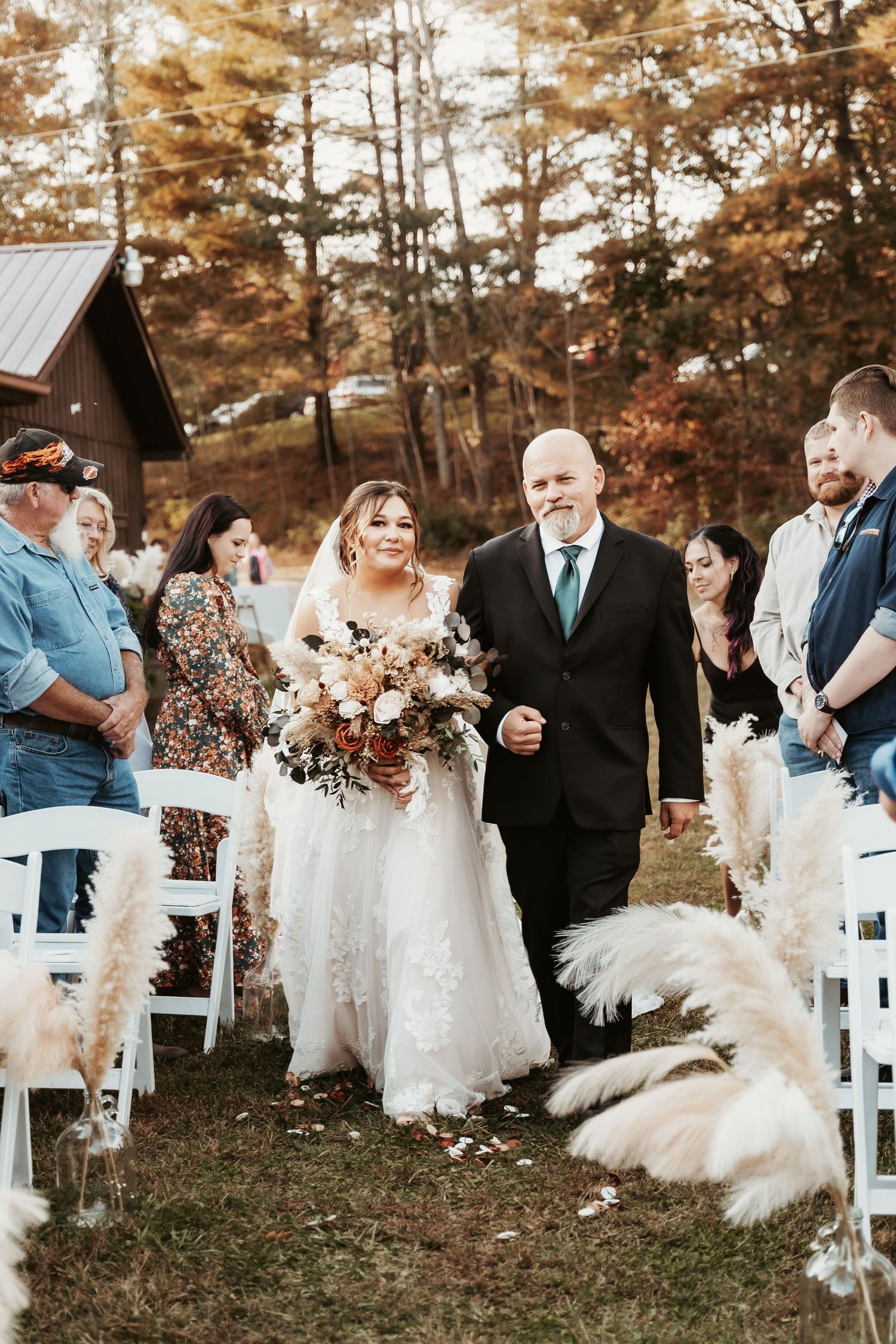 Outdoor autumn wedding ceremony with pampas grass decor and rustic cabin backdrop.