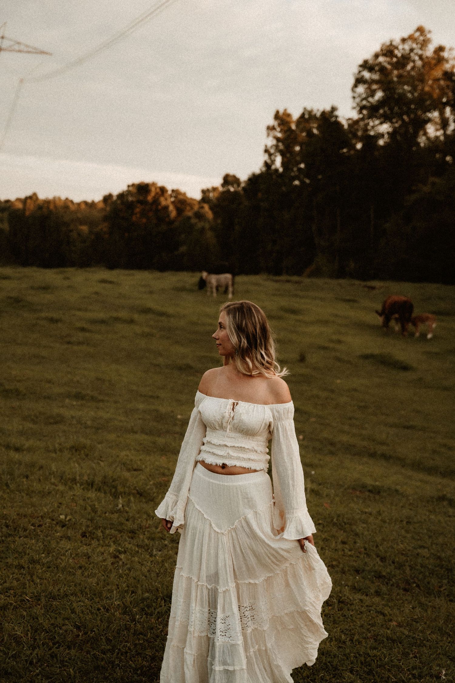 A romantic white bohemian dress flows in the breeze against a pastoral field backdrop with grazing horses.