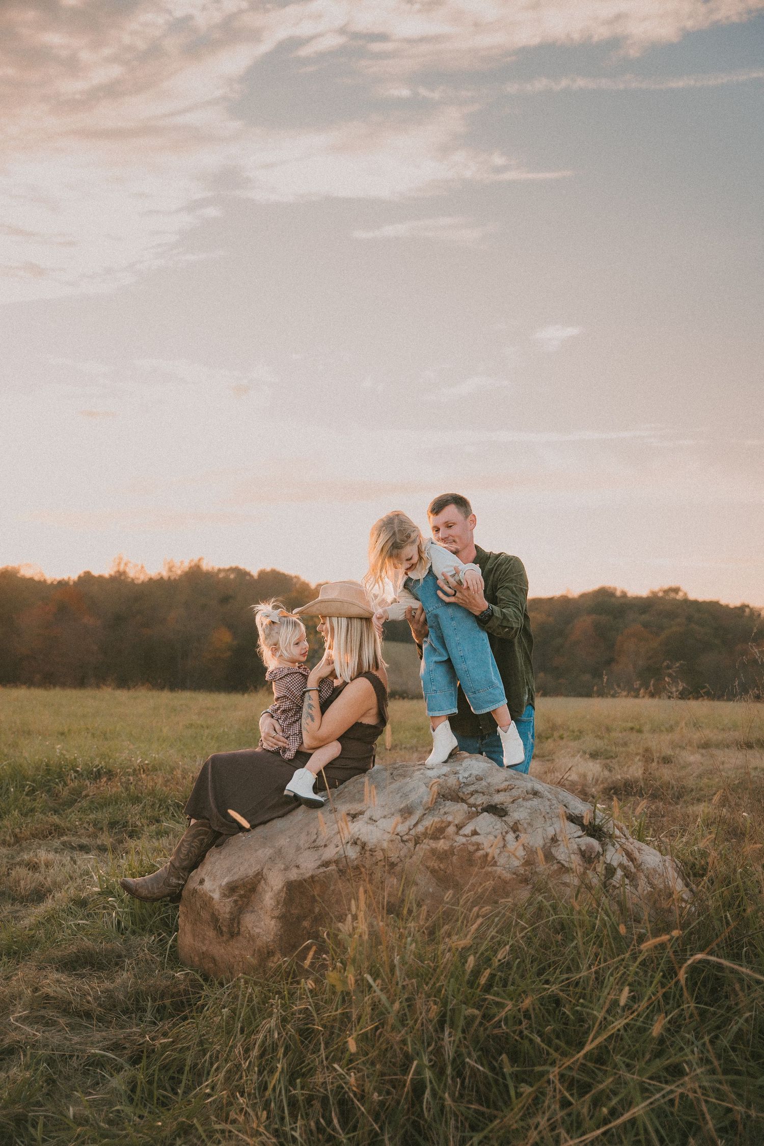 A couple sits together on a large rock in a grassy field at sunset.