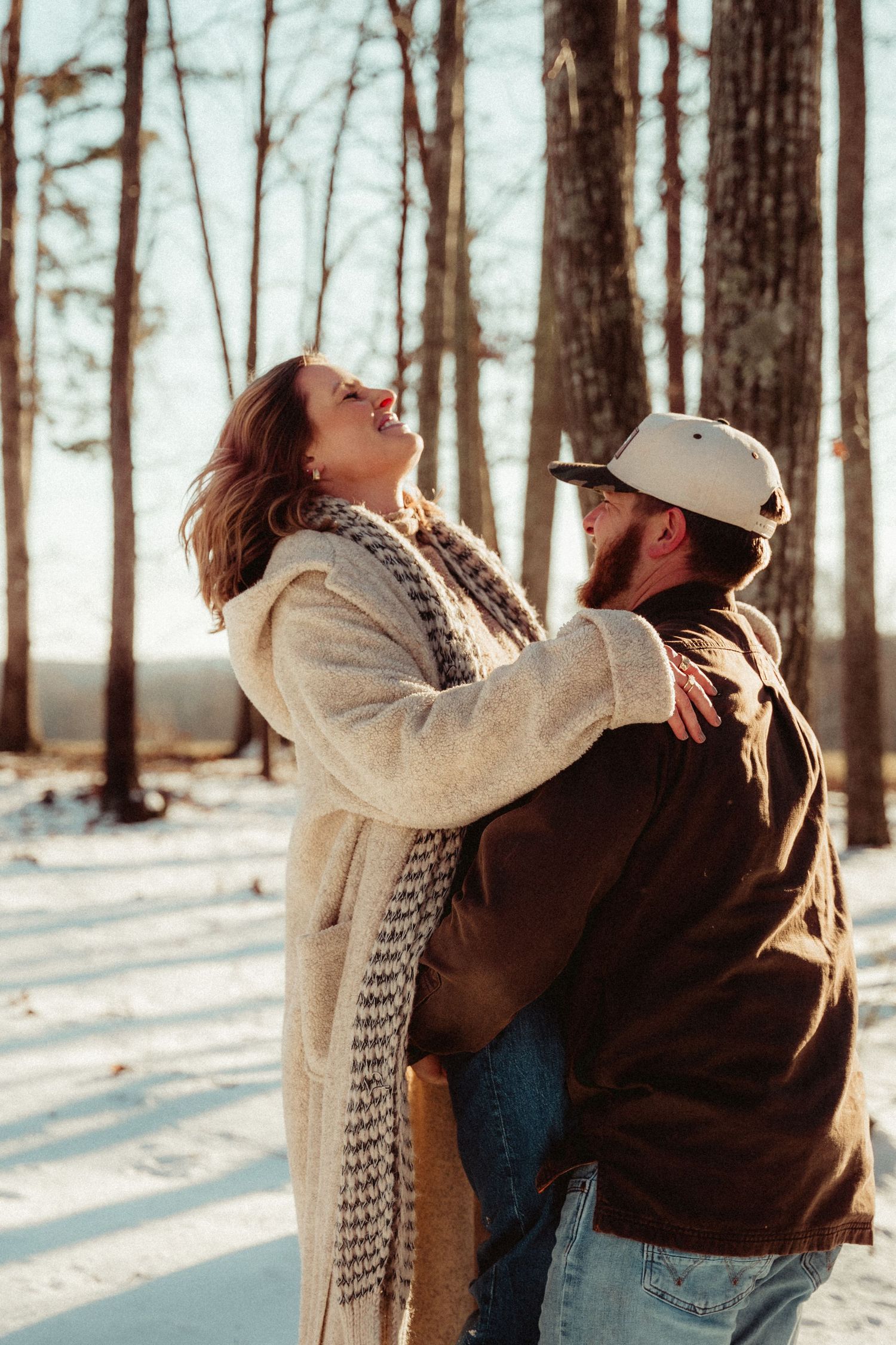A joyful couple embraces in a snowy forest setting with sunlight streaming through bare trees.