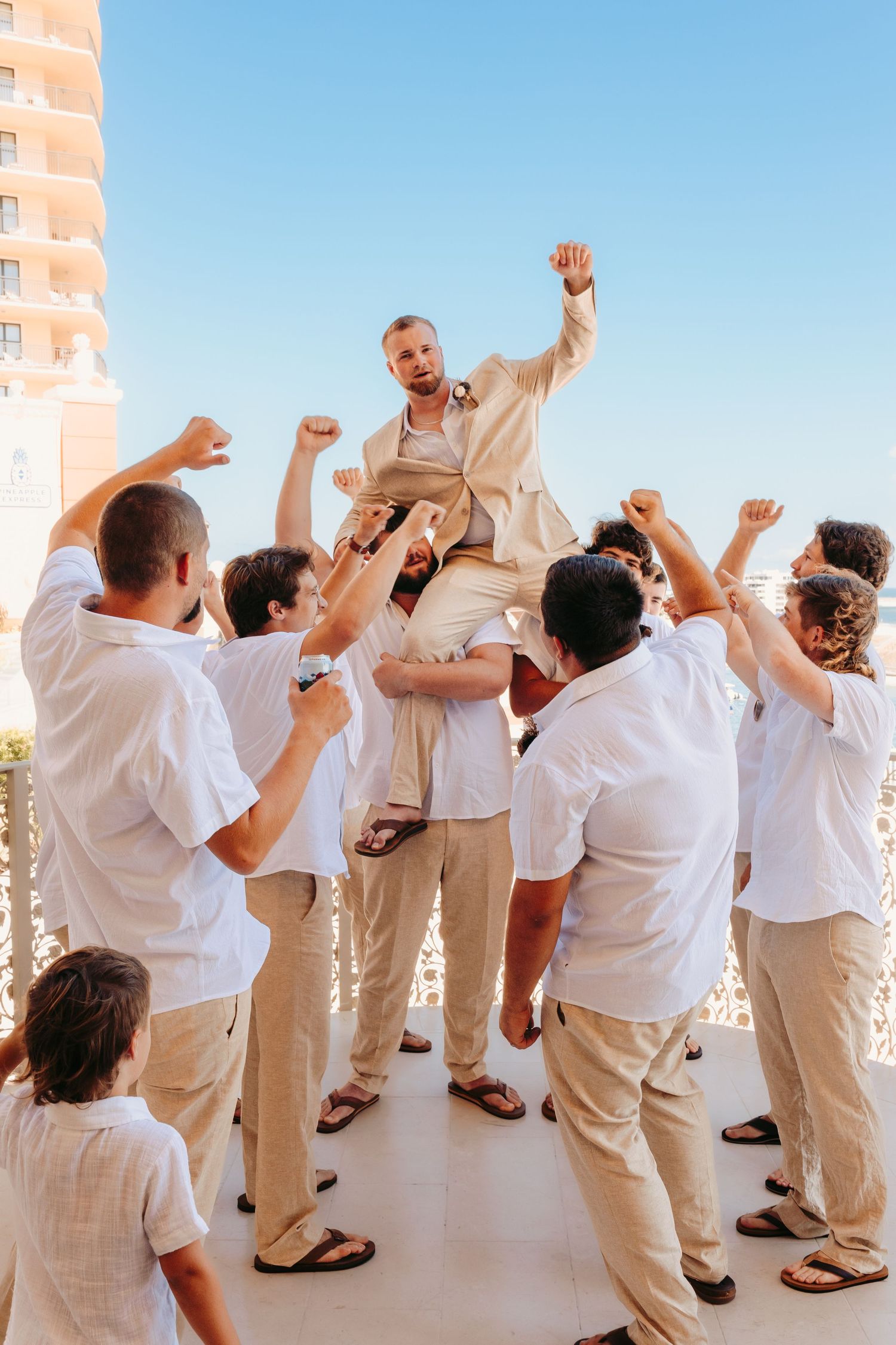 Wedding guests in white and beige attire celebrate joyfully with raised hands on a bright outdoor terrace.