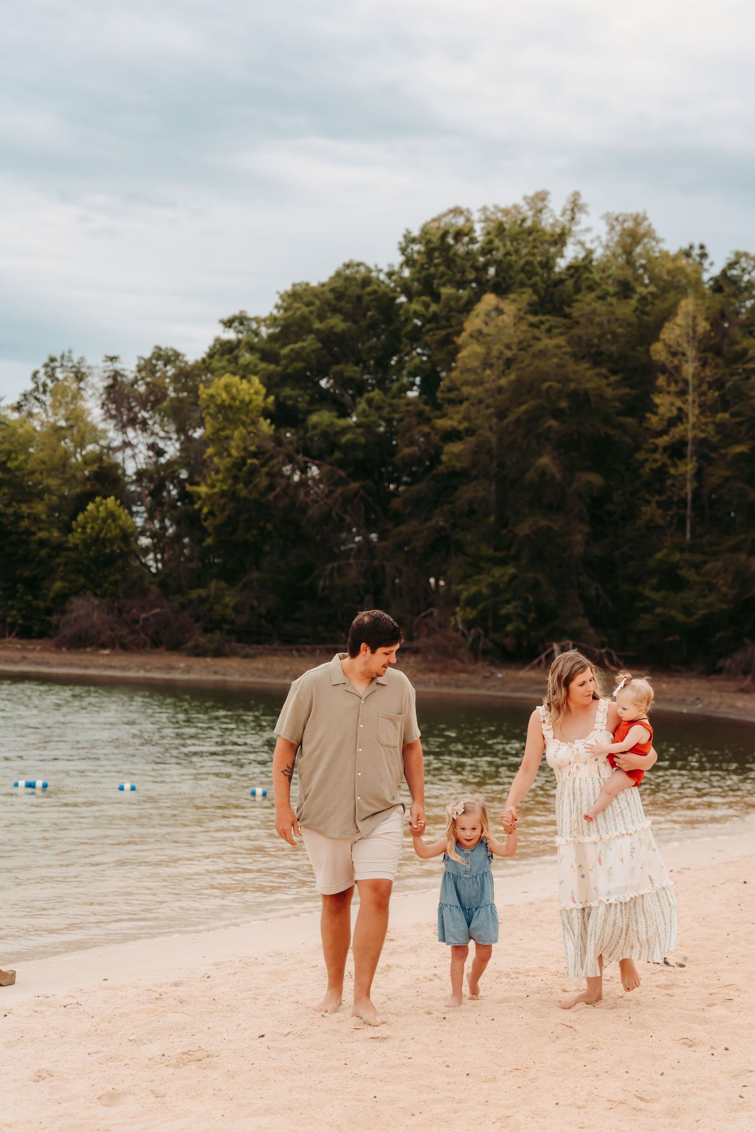 Family of four walking hand-in-hand on a sandy beach with lush green trees and calm water in the background.