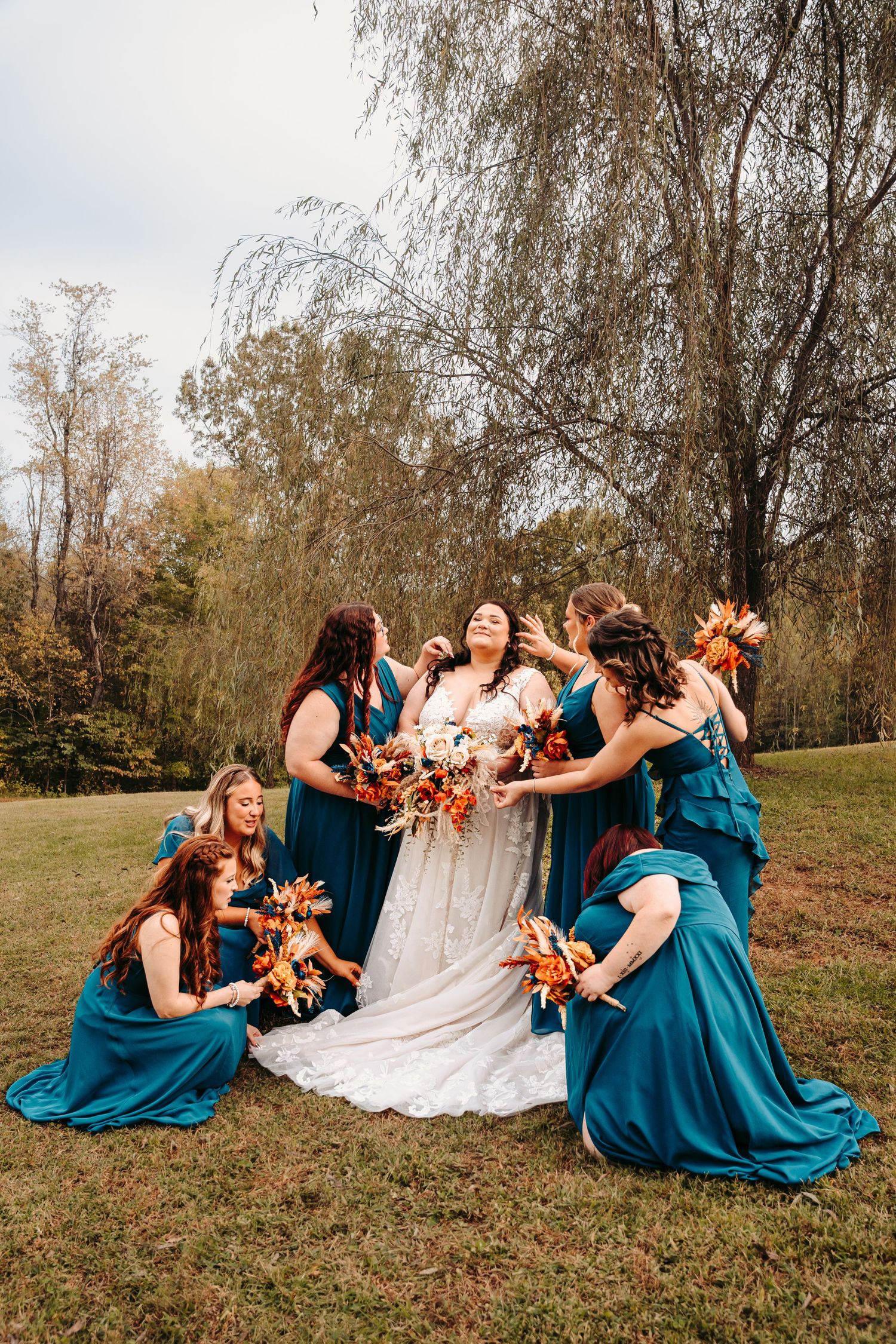 Wedding party in teal blue dresses laughs together while sitting on autumn grass.
