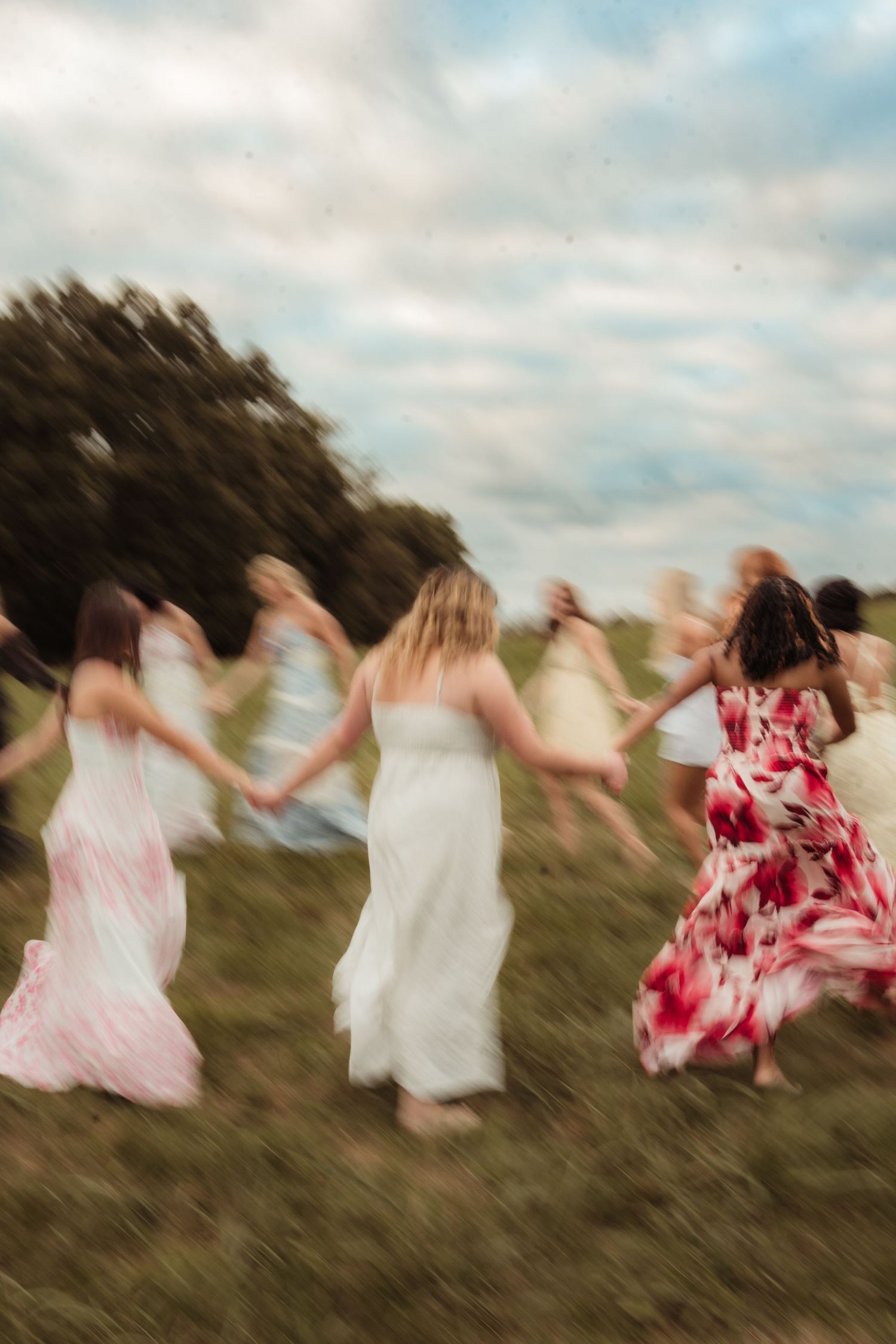 Group of women in colorful maxi dresses holding hands running through a green field under cloudy skies.