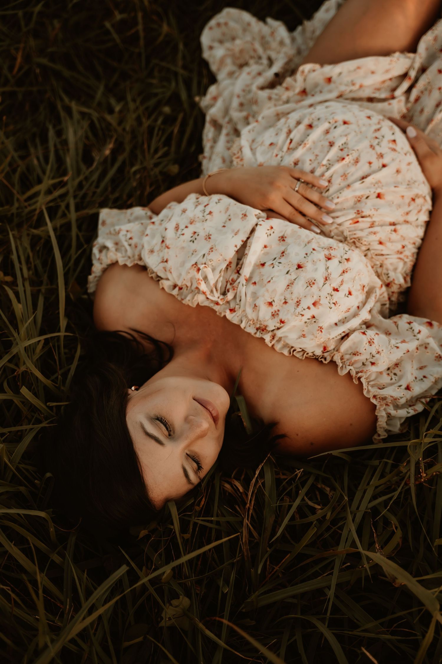 Artistic maternity portrait in floral dress lying in grass at dusk.