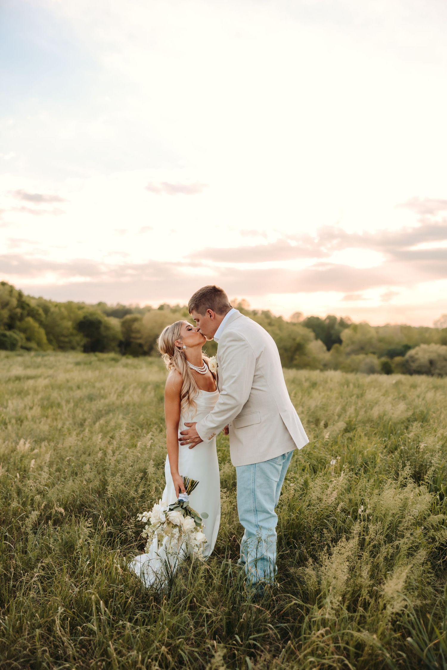 Bride and groom kissing in a golden meadow at sunset, bride holding white bouquet, groom in tan blazer.