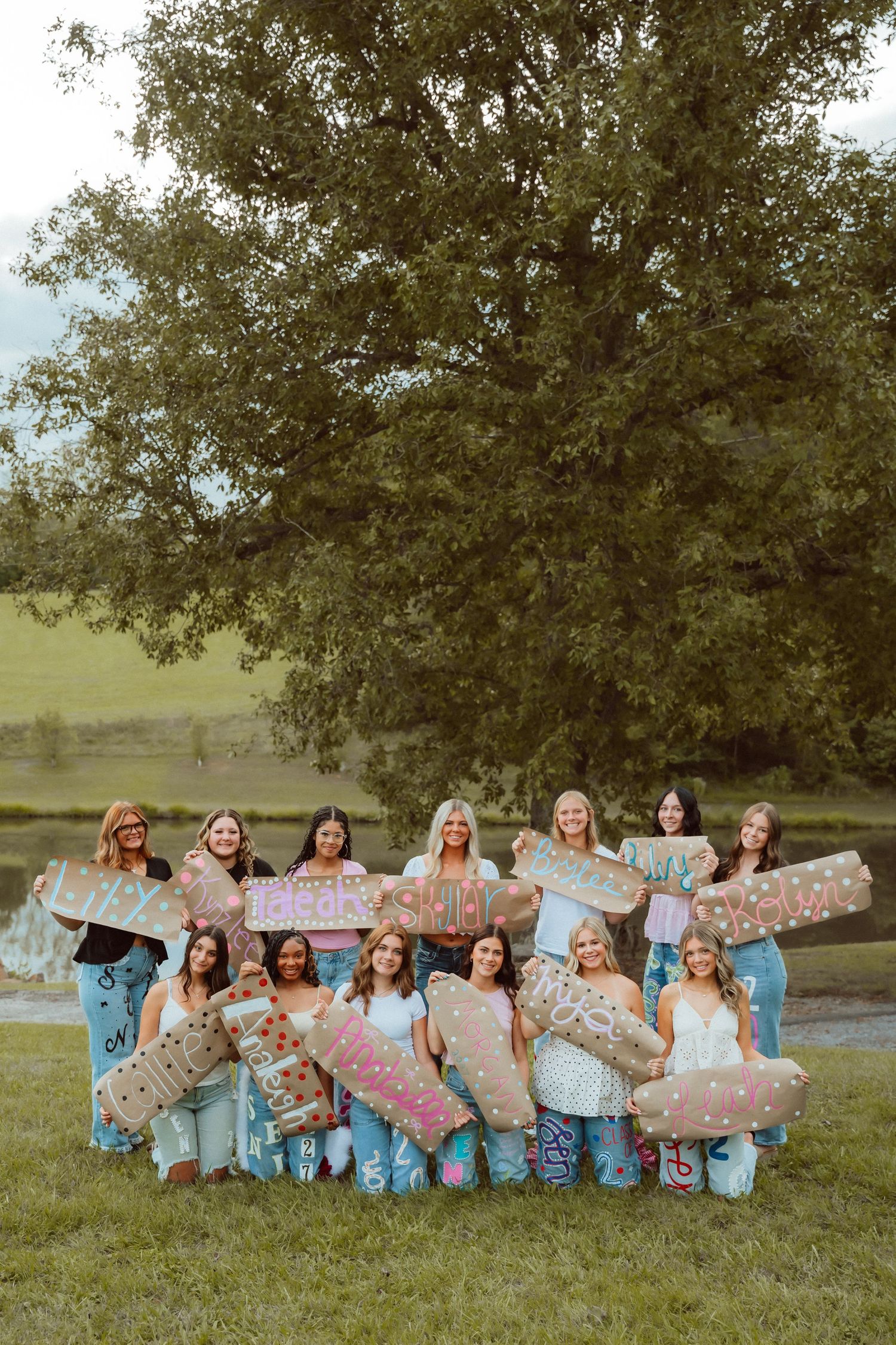 Group of women posing outdoors holding decorated signs near a large tree by a pond.