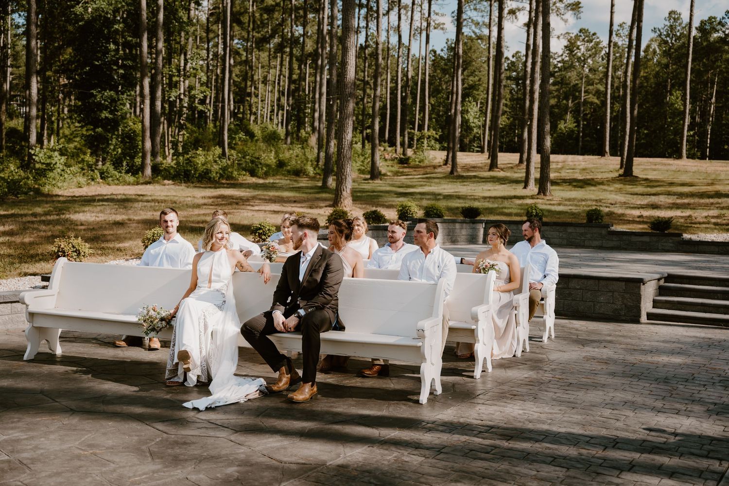 Wedding party seated on white chairs in pine forest setting.