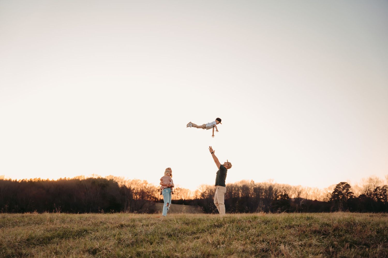 A family plays in a golden field at sunset, father tossing a child in the air while mother watches joyfully.